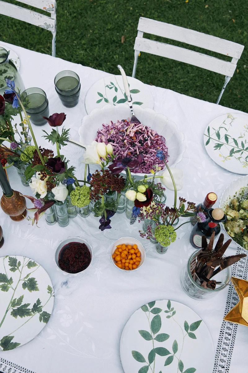 Outdoor table with floral plates, potted flowers, and colourful salad. Glasses and bowls on white tablecloth.