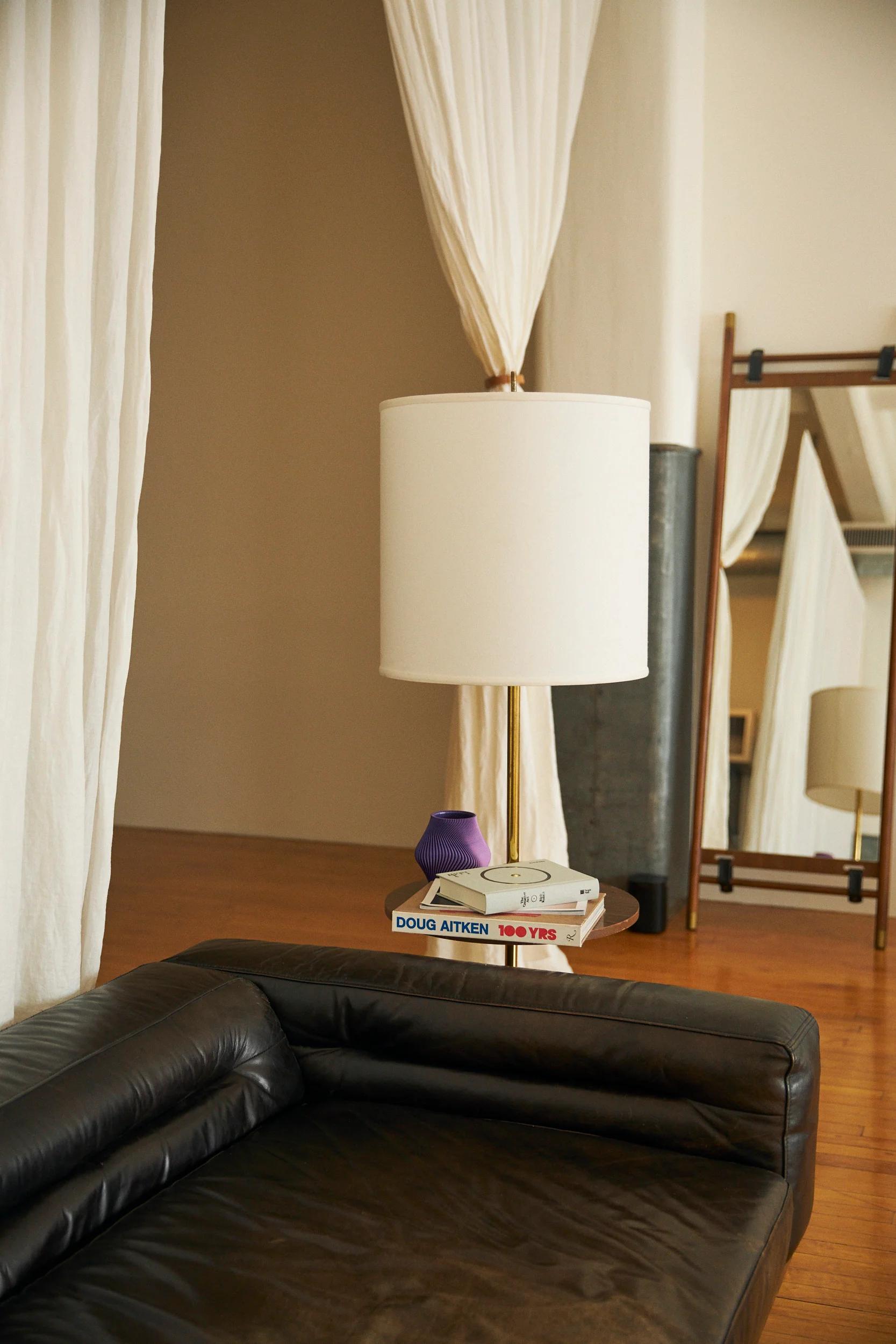 Modern living room with dark leather sofa, side table with books and cake kit. White lamp, purple ornament. Large mirror, white curtains, wooden flooring.