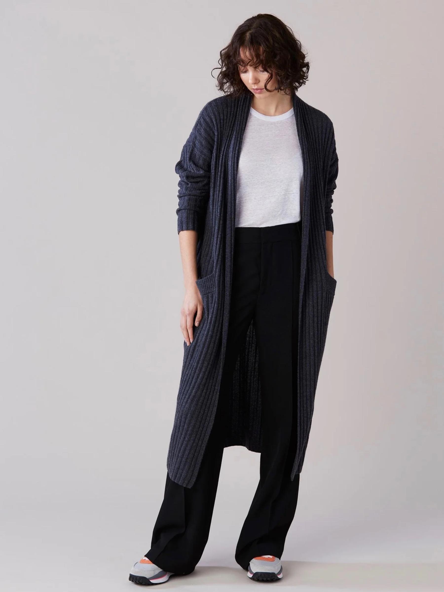 Wavy-haired person in long dark cardigan over white shirt and black pants. Standing, looking down, hands in pockets.