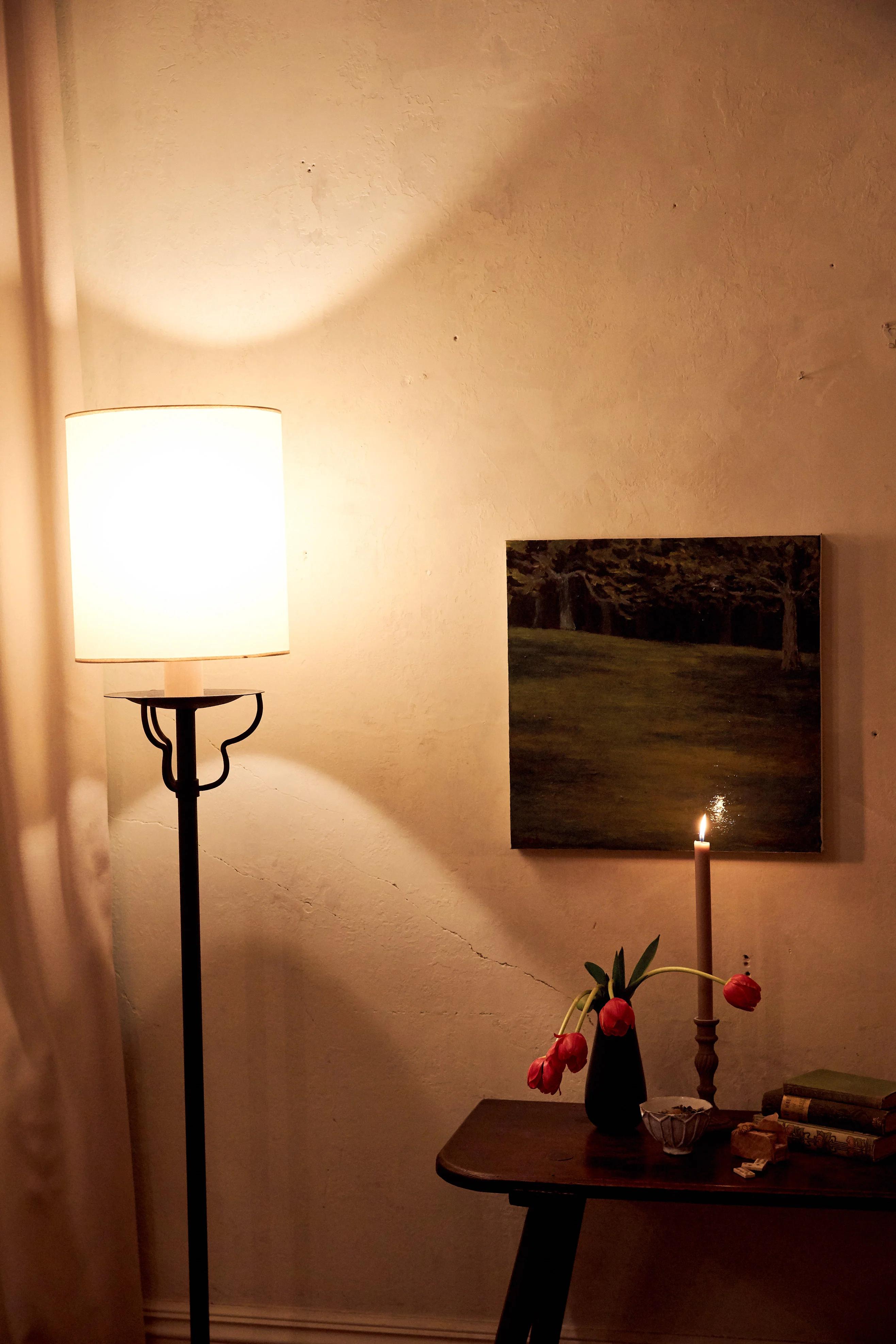 A standing lamp in the corner of a room glows across the walls. A small wooden table with flowers and a hanging picture are on the right.