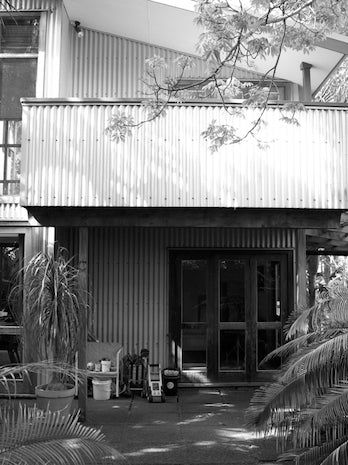 Black-and-white photo: Modern two-storey building with metal siding. Balcony above, glass doors below. Plants and furniture create a secluded feel.