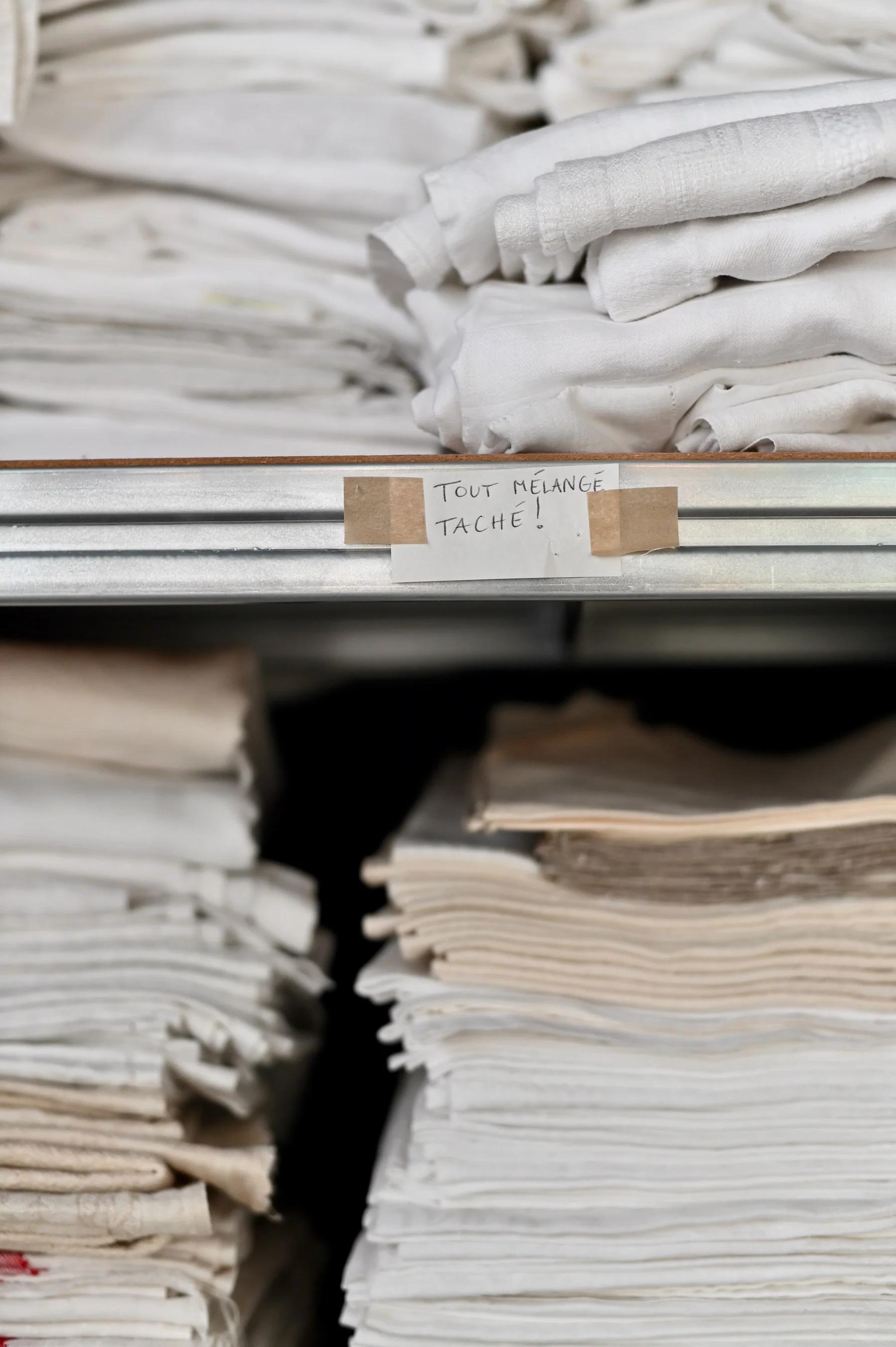 Stacked white and beige linens on metal shelves. Small handwritten French sign visible.