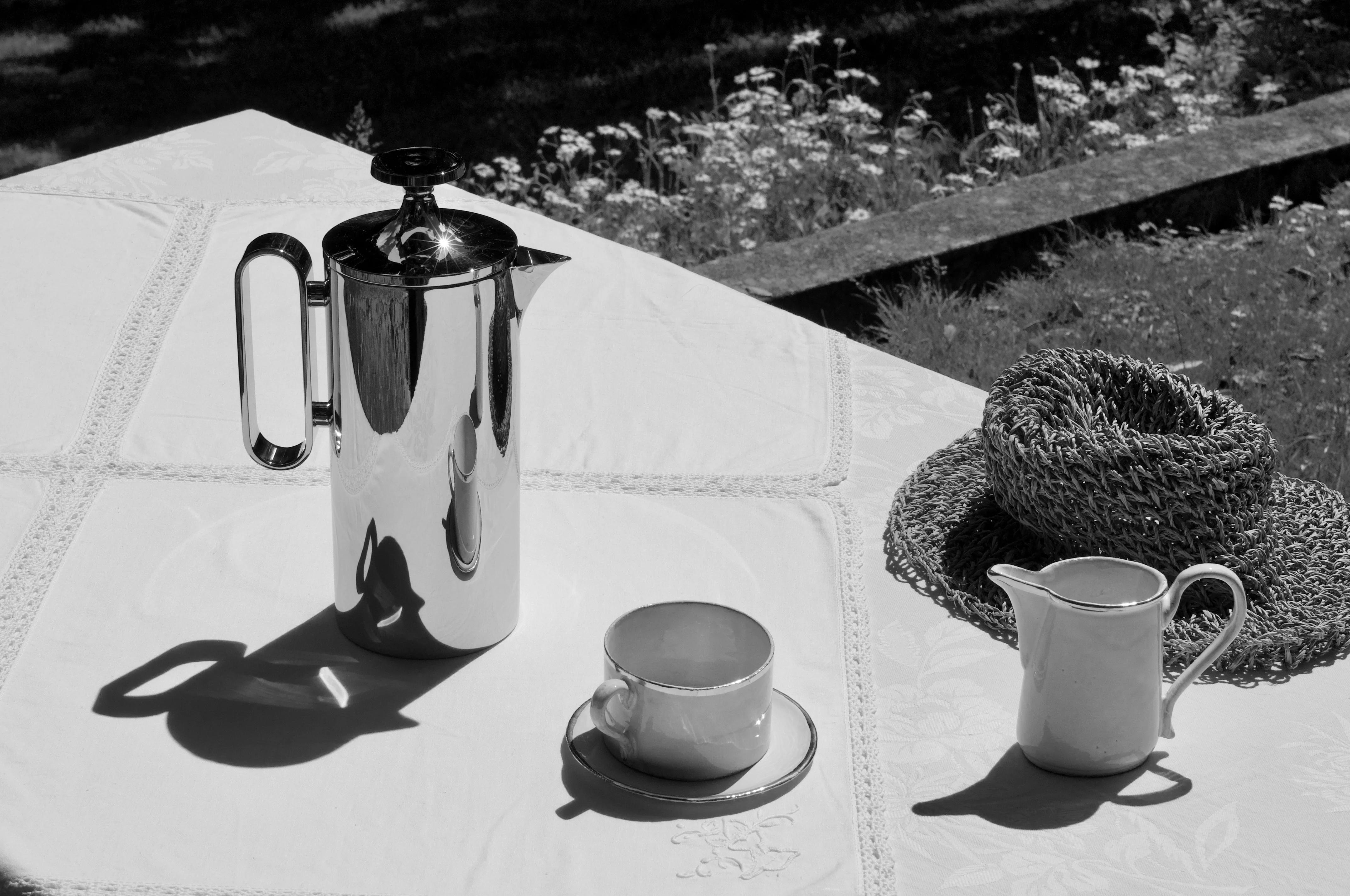 Black-and-white outdoor scene: Table with David Mellor cafetière, teacup, saucer, jug, and woven hat casting shadows. Stone ledge and sun-dappled foliage in background.