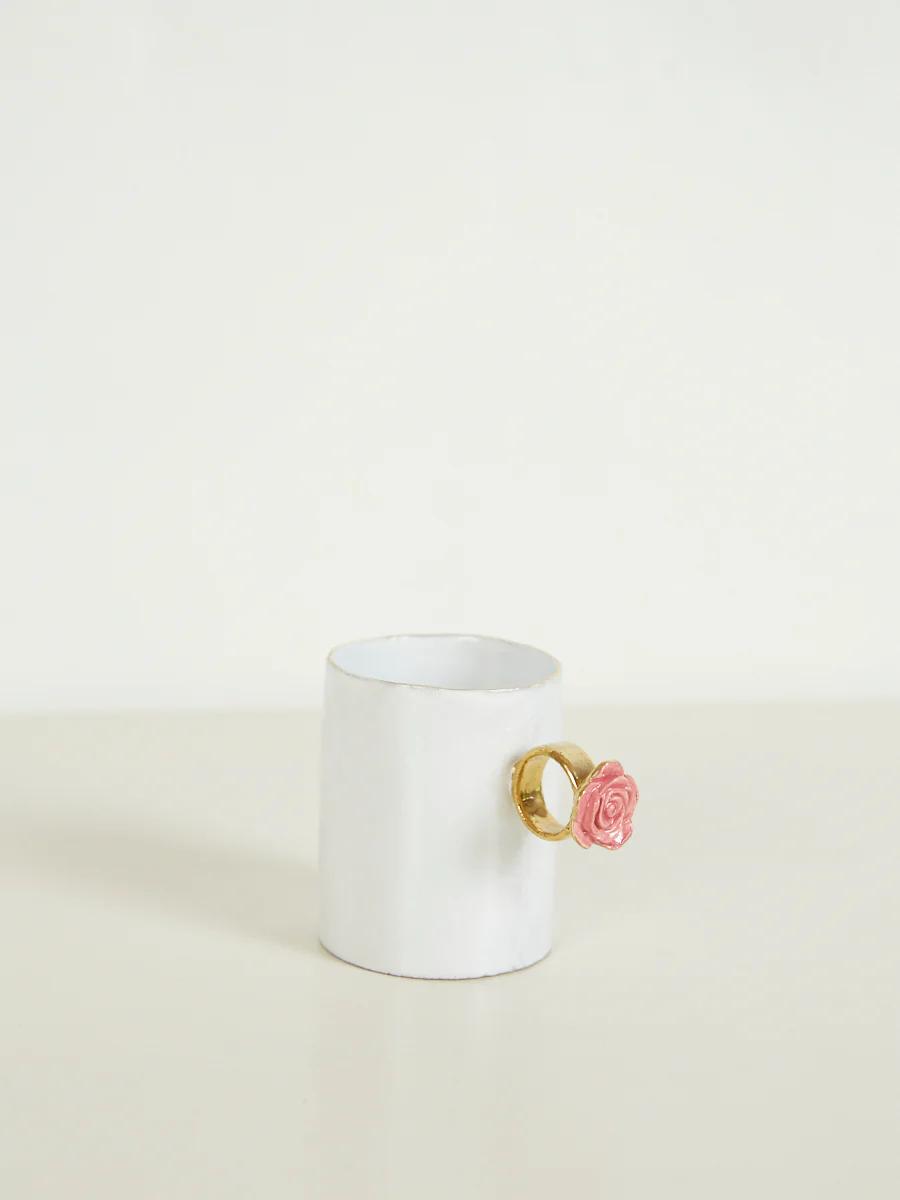 White ceramic mug with handle resembling pink rose in gold ring. Plain, light-coloured background.