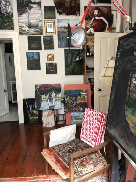 Cozy studio with wooden floors. Paintings on walls. Chair near easel with unfinished canvas. Max Thomson's art supplies around.