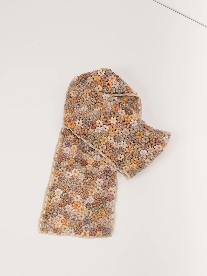 A flat-lay of a floral crochet scarf in shades of beige, brown, white, and pink. Designed by Sophie Digard, the scarf features intricate hand-crocheted flowers.