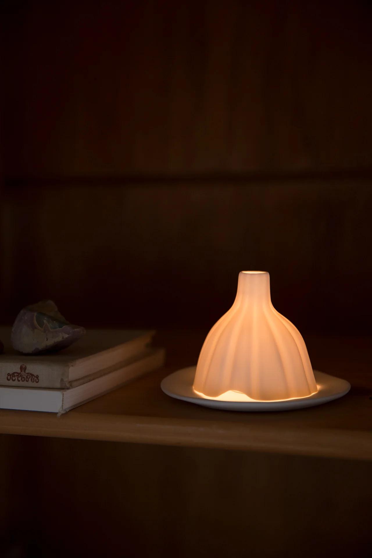 Glowing table lamp with bell-shaped shade on wooden surface. Stack of books and fabric object nearby. Dimly lit background.