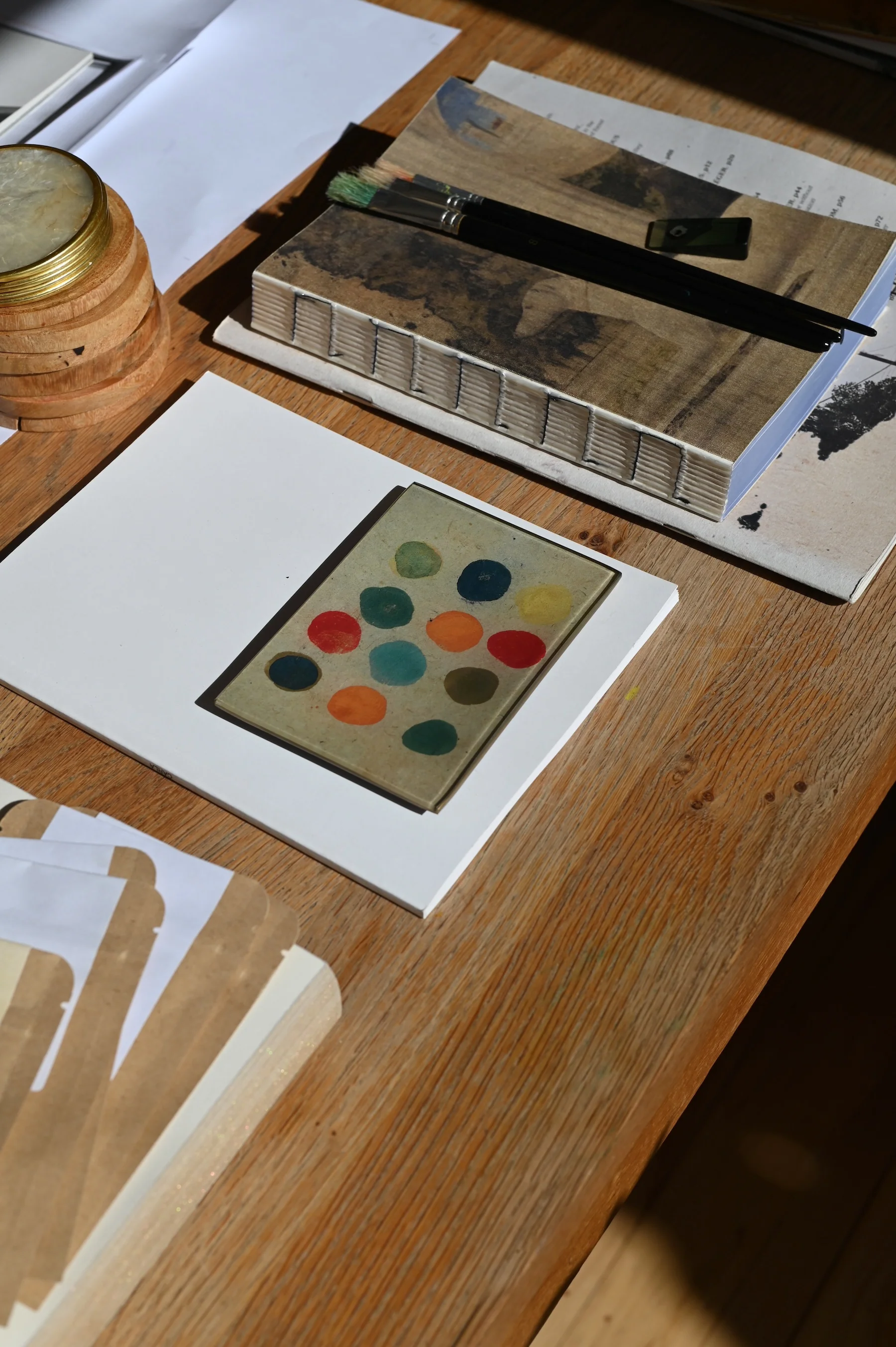 Wooden table with art supplies, paint samples, cards, and découpage glassware. Neatly arranged papers.