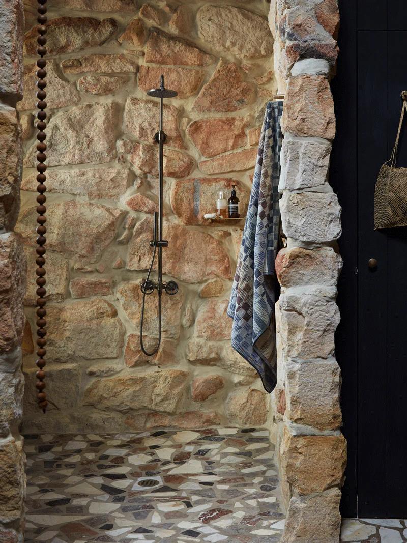 A rustic shower with stone walls and a black shower head, featuring a grey and blue towel hanging from a hook next to shower essentials on a shelf.