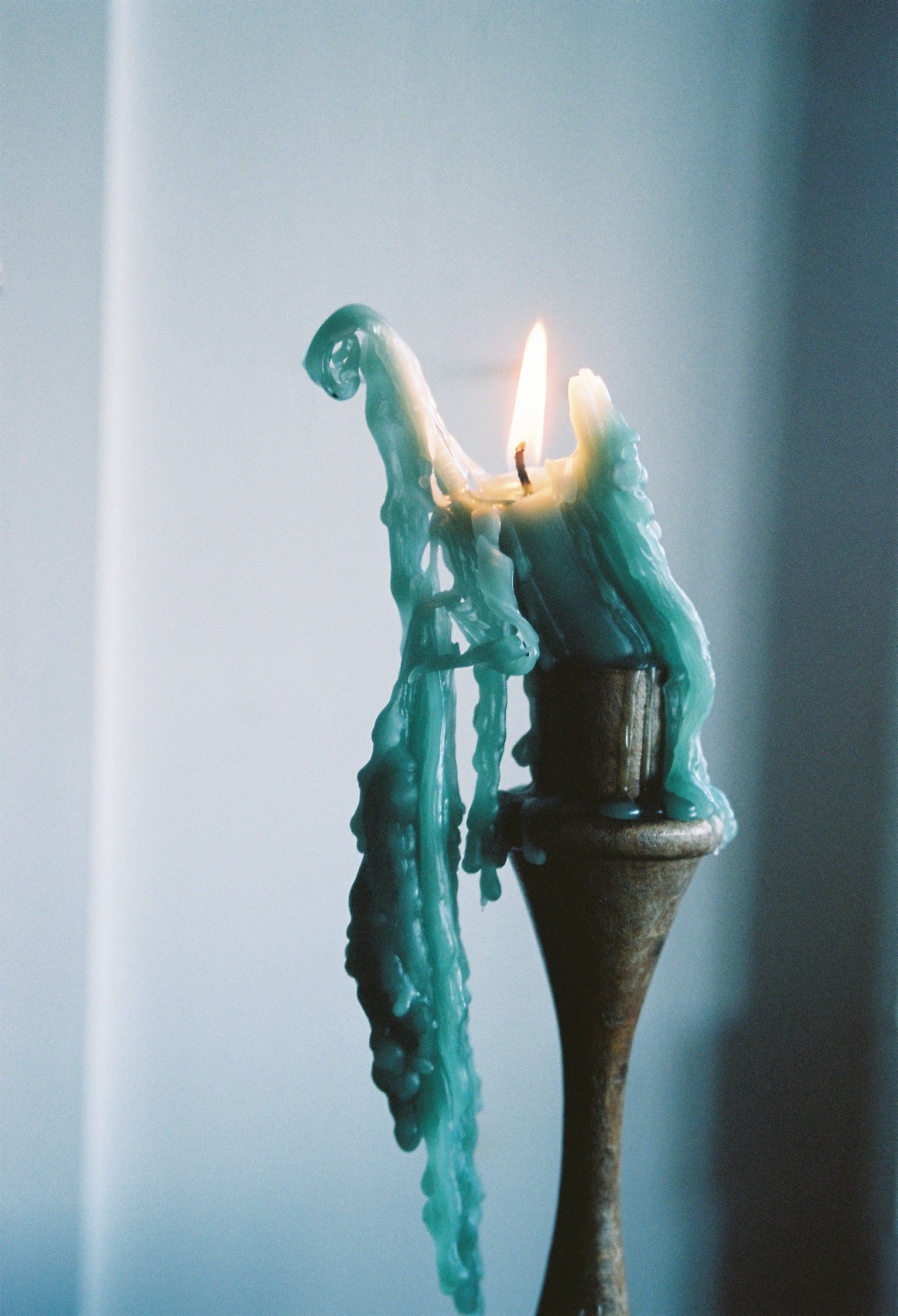 A light blue candle burning on a wooden holder, with wax dripping down the sides, creating a serene and tranquil atmosphere by the ocean.