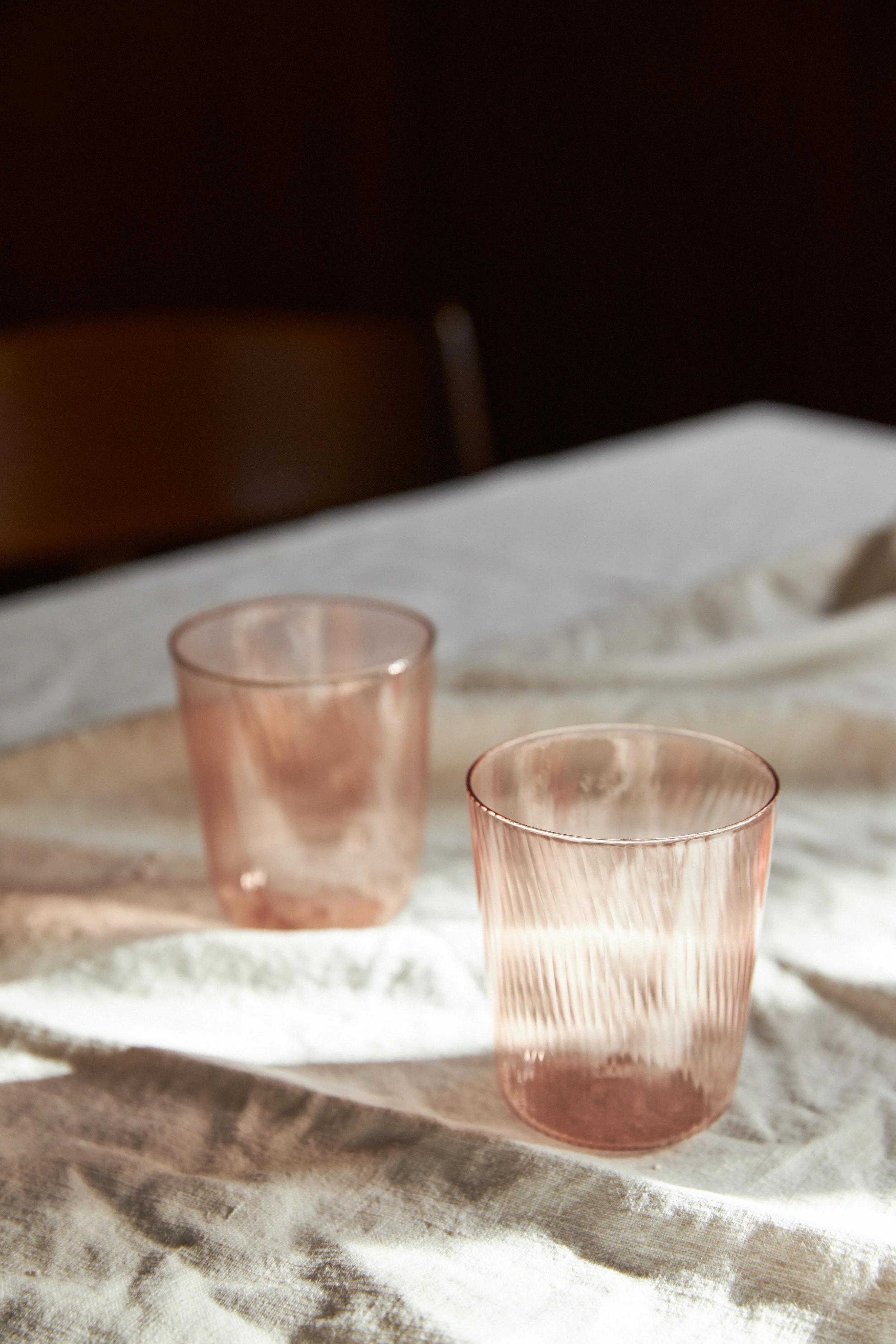 Two ribbed, translucent pink glasses on a white tablecloth. Sunlight creates shadows and reflections of the glasses on the tablecloth.