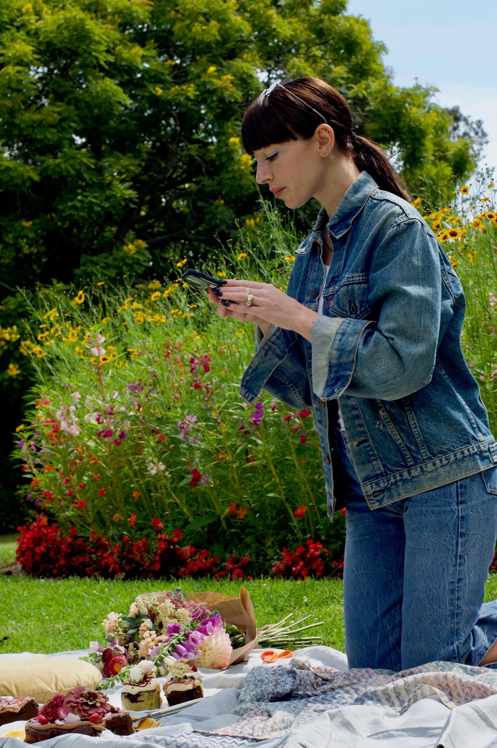 A woman kneels to photograph a picnic spread in Auckland, featuring treats from The Caker bakery. The scene is surrounded by flowers and lush greenery.
