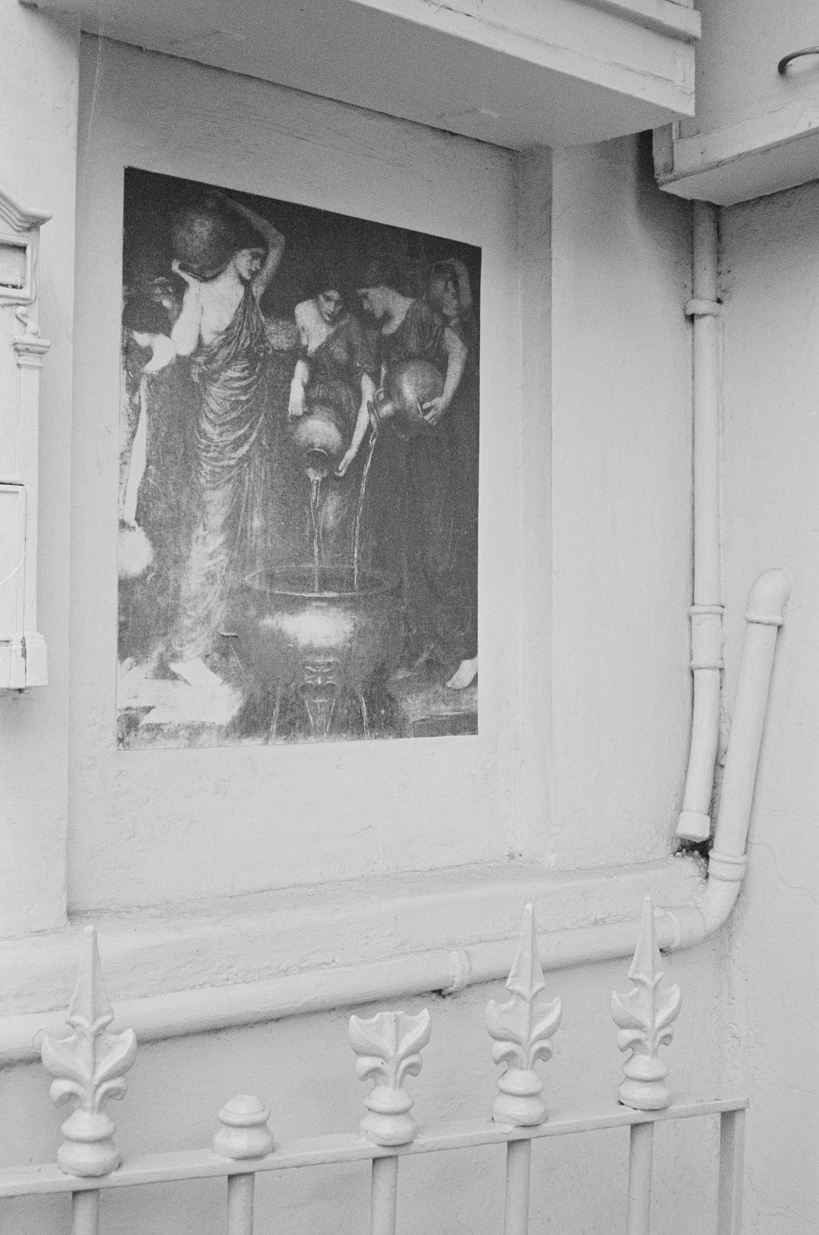black and white image of poster of maidens and water bowl outside a home's gate