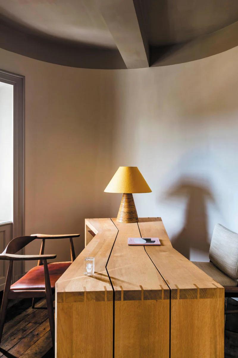 A minimalist office with a geometric wooden desk holding a yellow lamp and notebook. Two chairs nearby complete the French-inspired design.