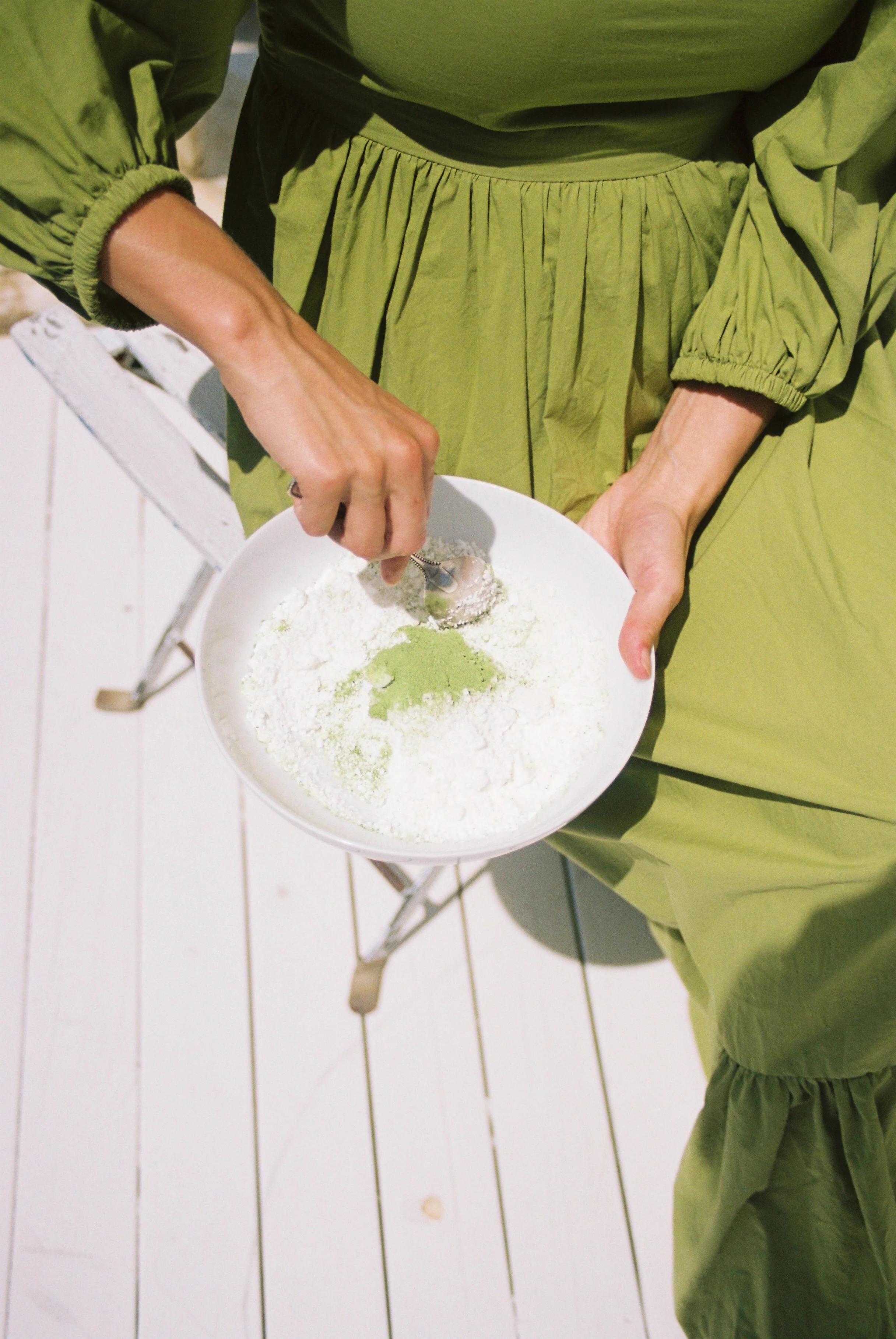 Person in green dress mixing green powder with flour in white bowl. White slatted wooden background.