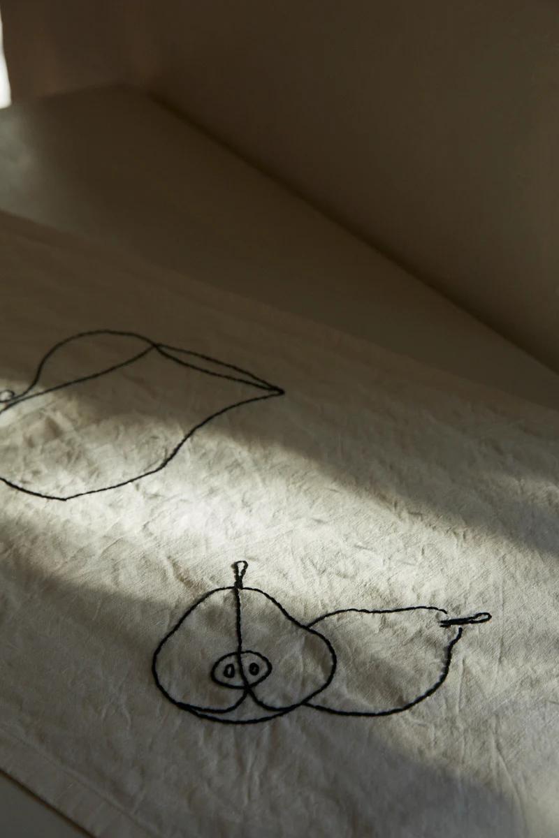 Fabric with black line drawings of two apples, one whole and one halved. Soft natural light creating subtle shadows.