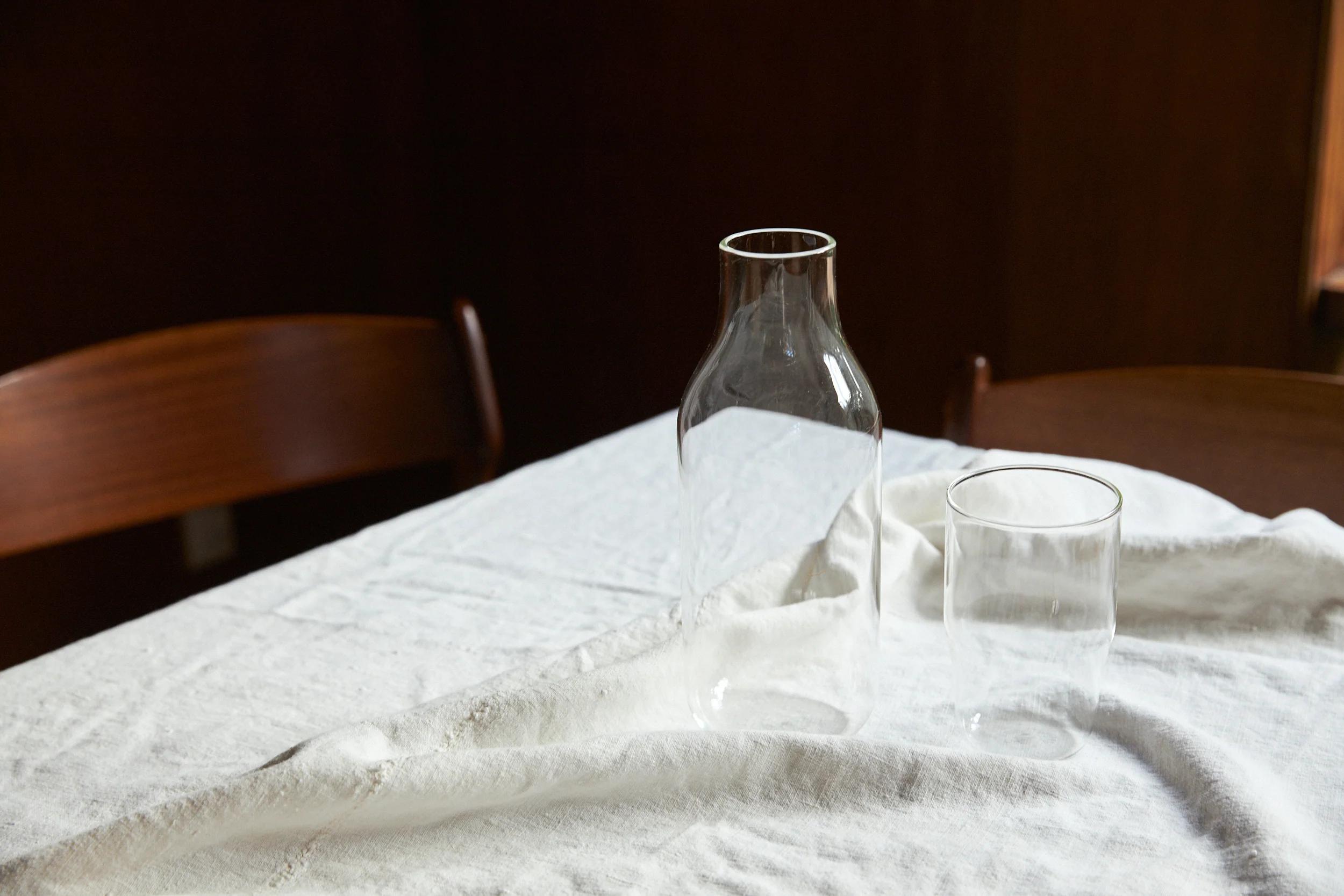 A glass carafe and drinking glass sit on a white tablecloth on a wooden table. Two wooden chairs are placed around the table.