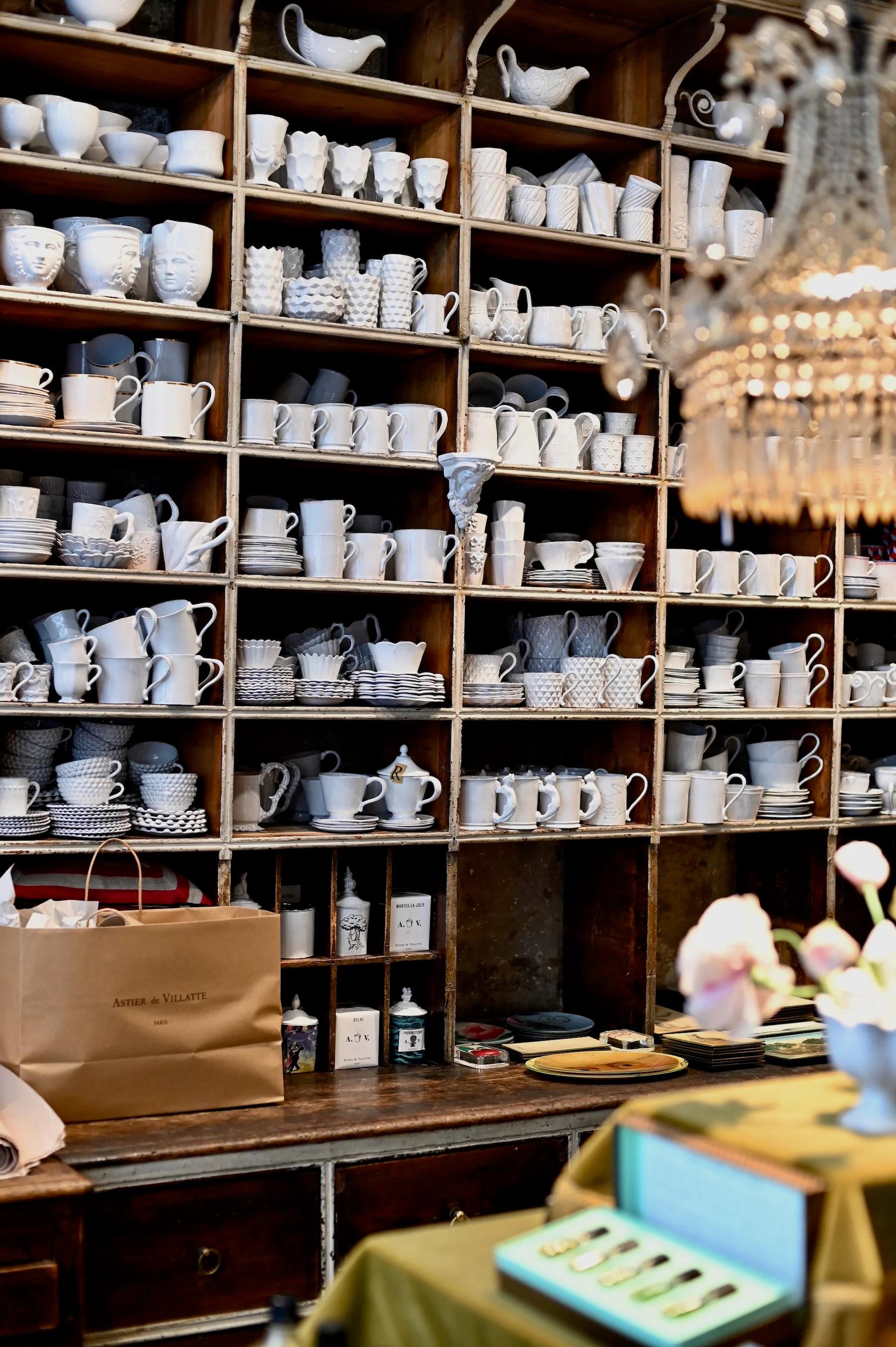 Ceramic dishware on wooden shelves. Teapots, cups, saucers, and plates. Bags and decorative items below. Soft lighting and chandelier.