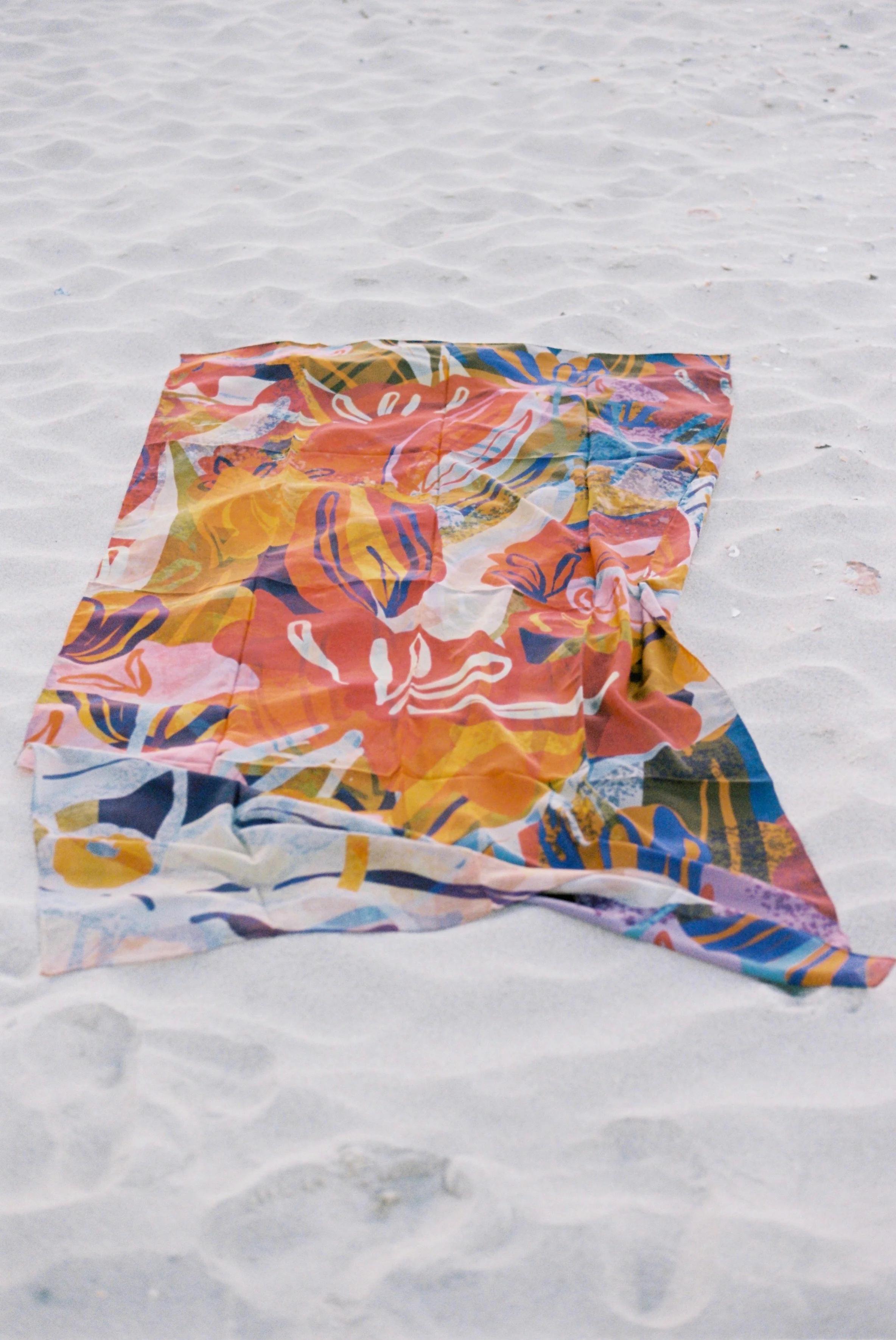 Colourful blanket with tropical patterns on sandy beach. Orange, pink, green, and blue hues with floral designs.