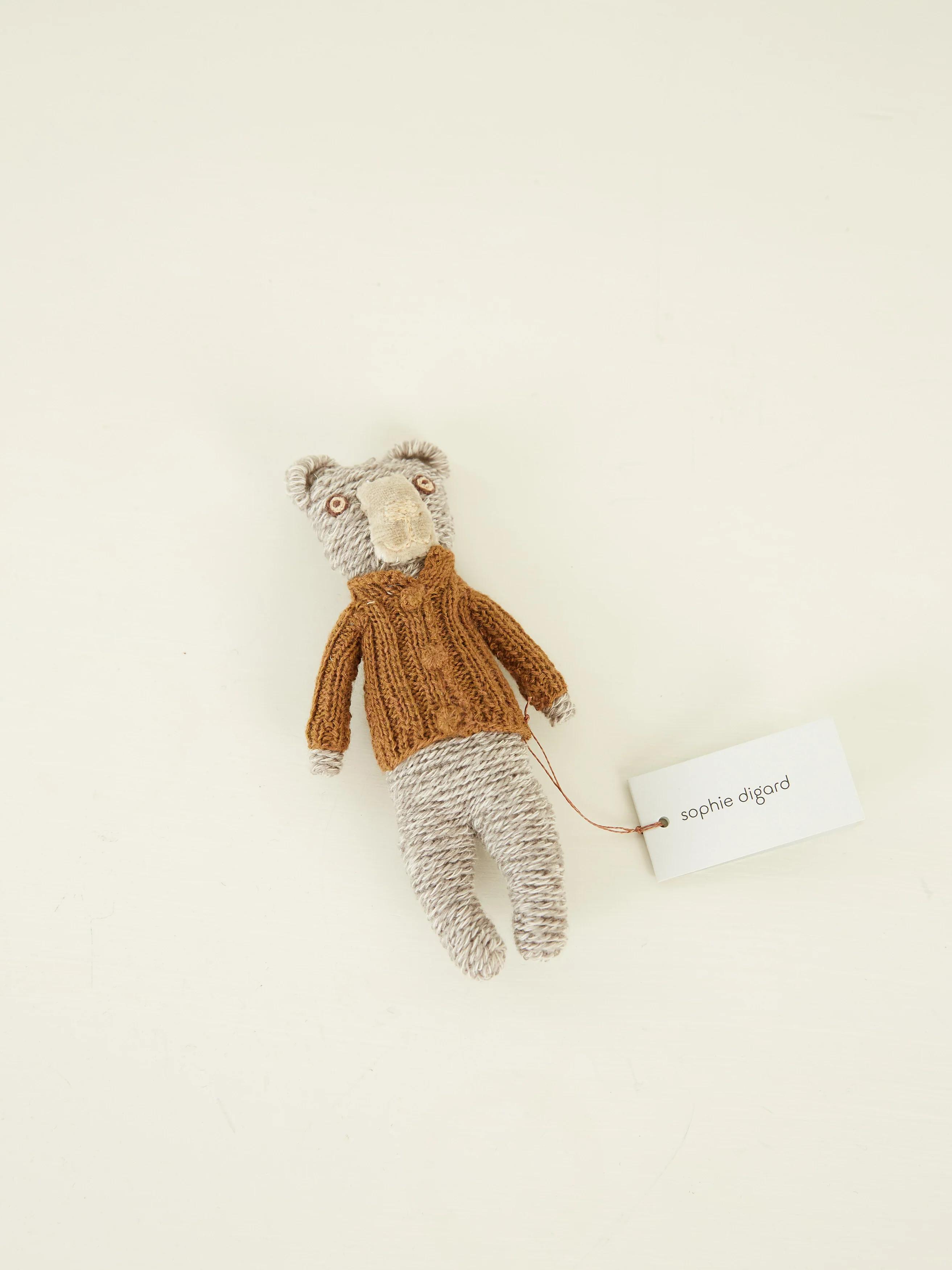 A small grey bear toy wearing a brown sweater with a “Sophie Digard” tag, lying flat on a light surface.Javier Bear, 15cm