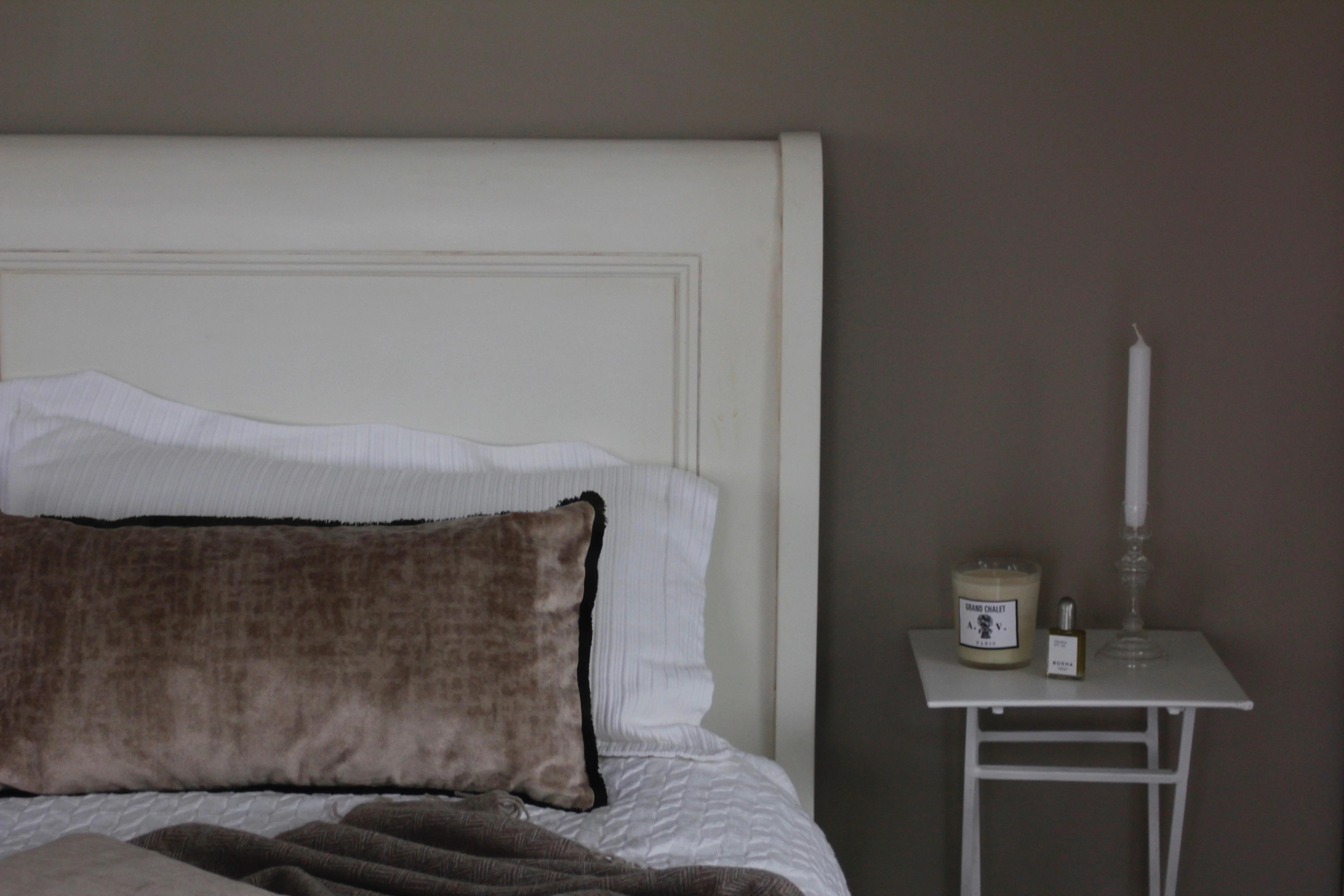 White bed with brown pillow. Nightstand holds Astier de Villatte candle, perfume, and tall candlestick. Soft brown walls complete cosy winter scene.