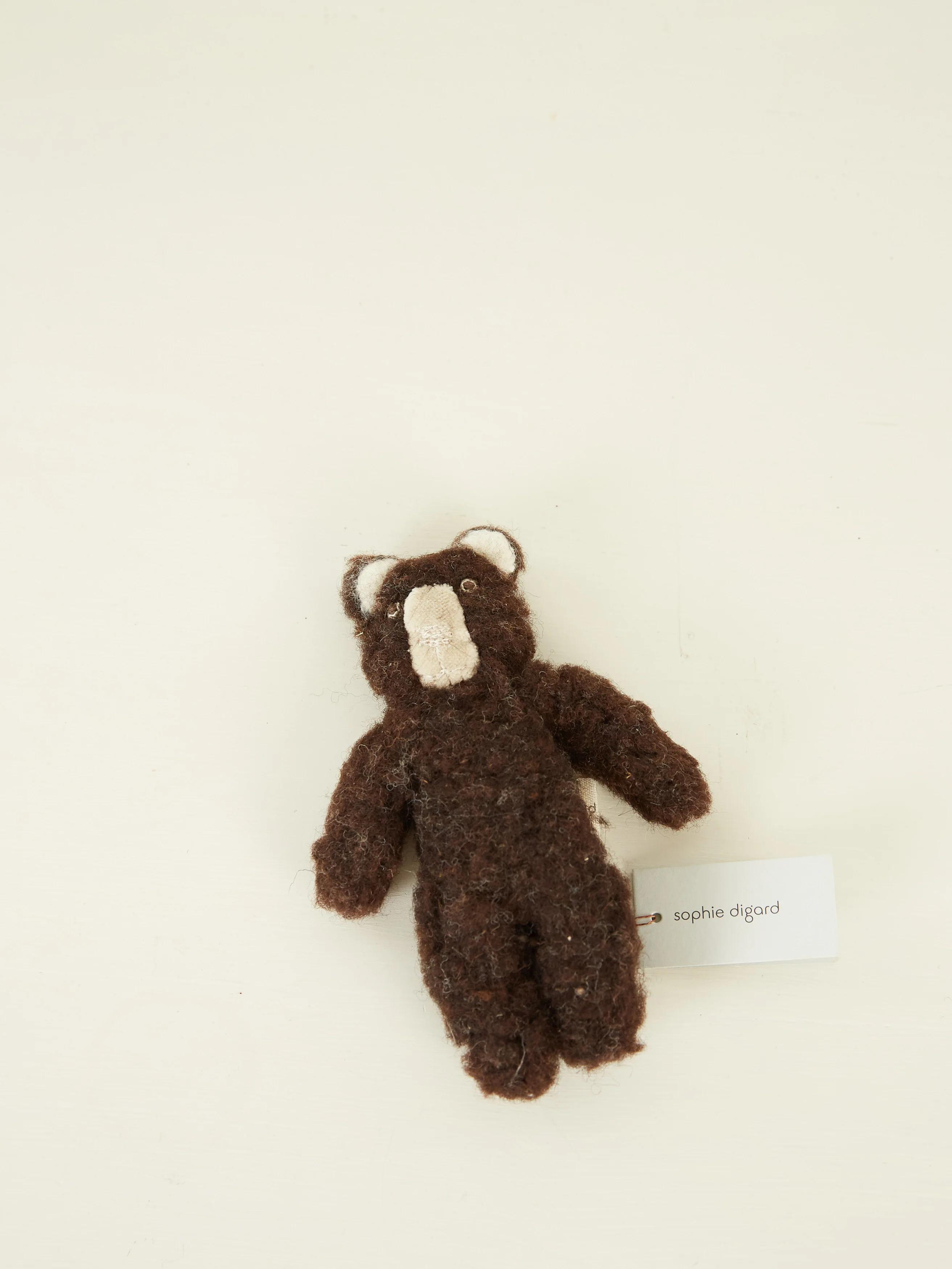 A small brown teddy bear with a white snout and ears, lying on its back with a “Sophie Digard” tag on a light surface.