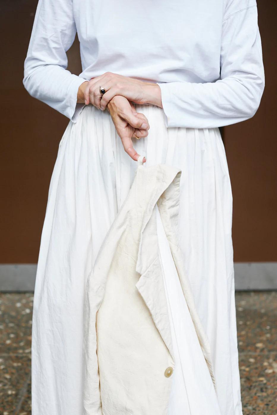 Person in white top and skirt holding beige jacket. One hand touches wrist. Brown wall background.