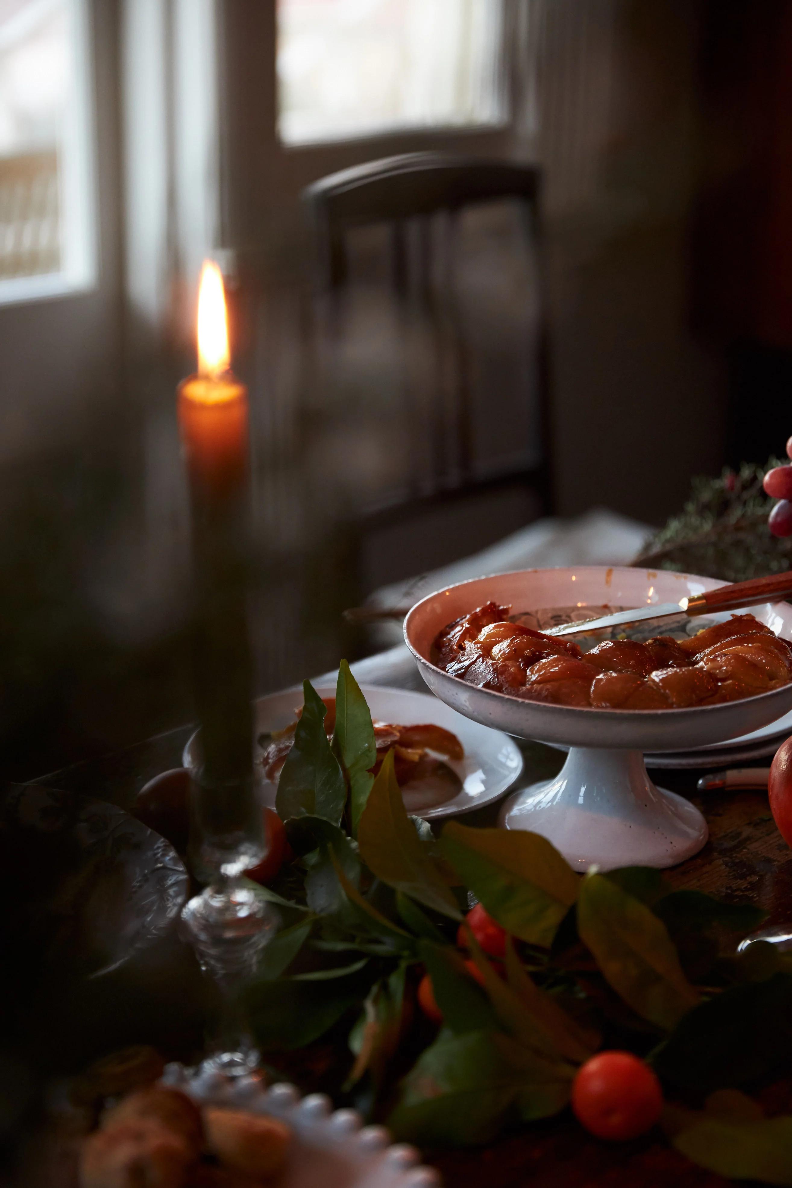 Cosy indoor scene with lit candle and homemade Fruit Tarte Tatin on white pedestal plate. Fresh apples and pears surround the tart, creating festive atmosphere by window.