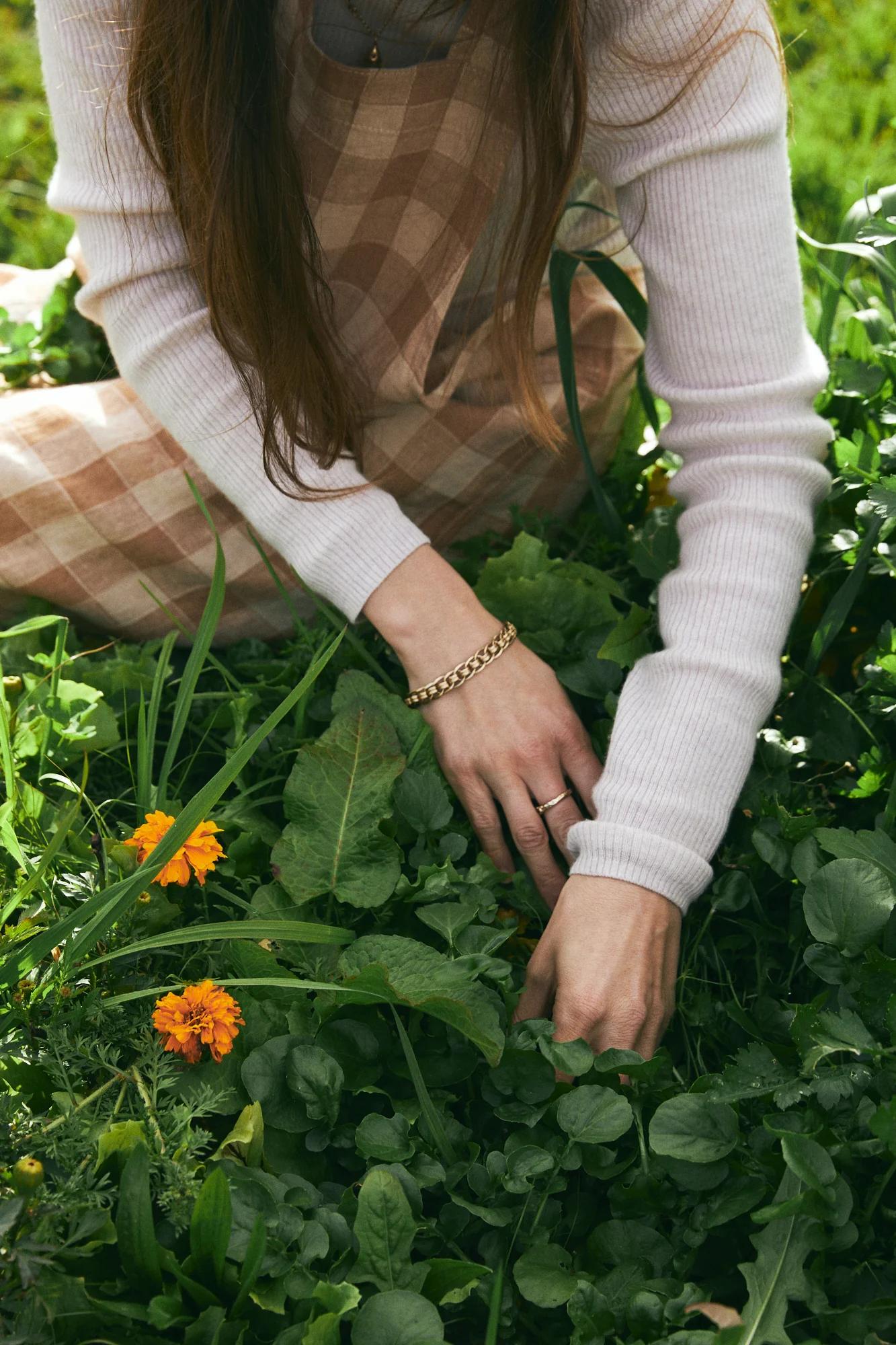 Person in checkered dress gardening. Holding greenery amid green leaves and orange flowers.