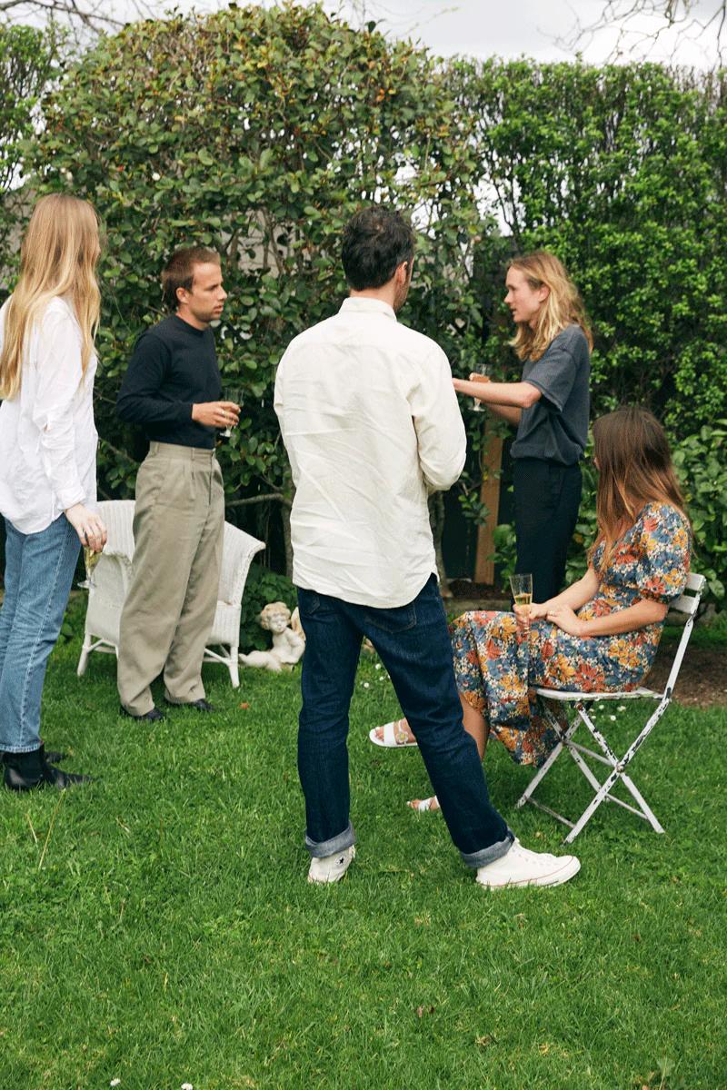 Five people chat in garden during holiday season. Two seated, three standing. Lush green bushes and trees in background.