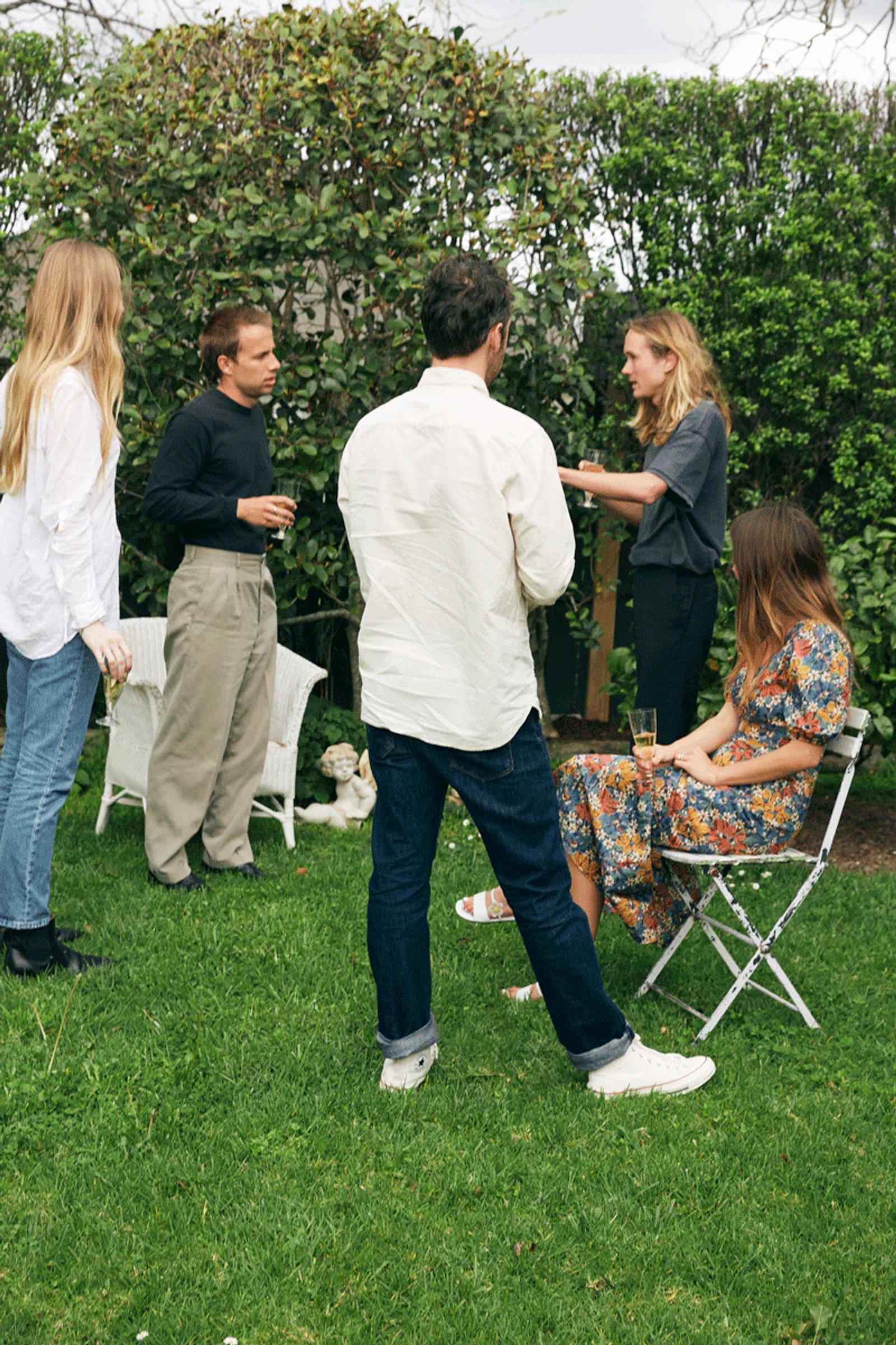 Five people chat in garden during holiday season. Two seated, three standing. Lush green bushes and trees in background.