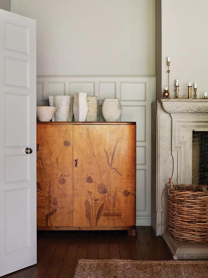 Cozy interior with floral wooden cabinet and ceramic vases. Stone fireplace with brass candleholders and nearby wicker basket. Wooden floors and paneled walls.