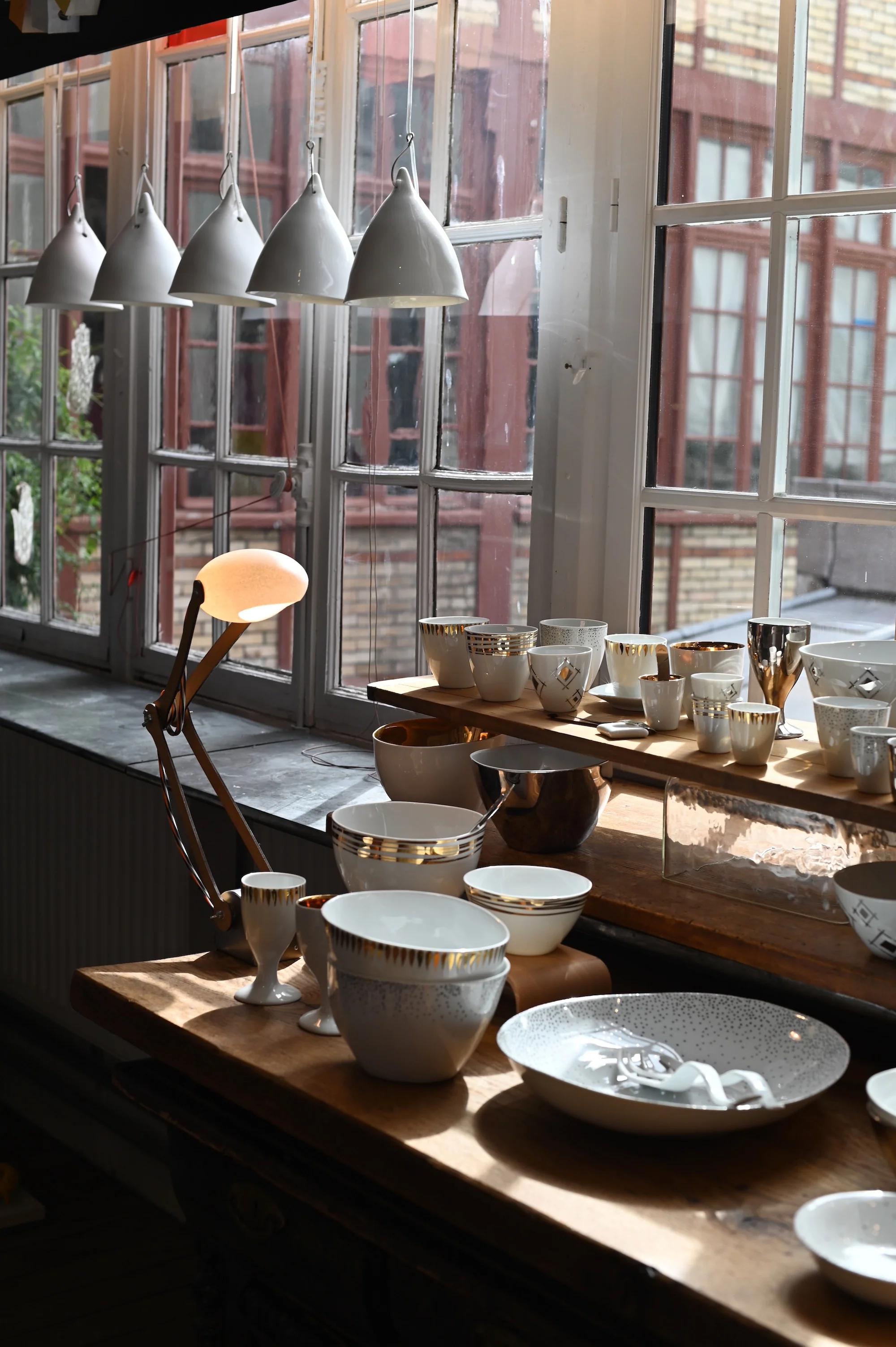 Sunlit table by window with white and gold ceramics. Modern lamp. View of brick buildings and greenery outside.