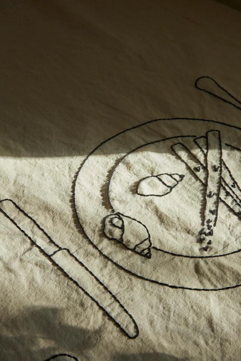 Fabric with black thread embroidery of knife, fork, plate with pastries, and breadsticks. Sunlight shadow visible.