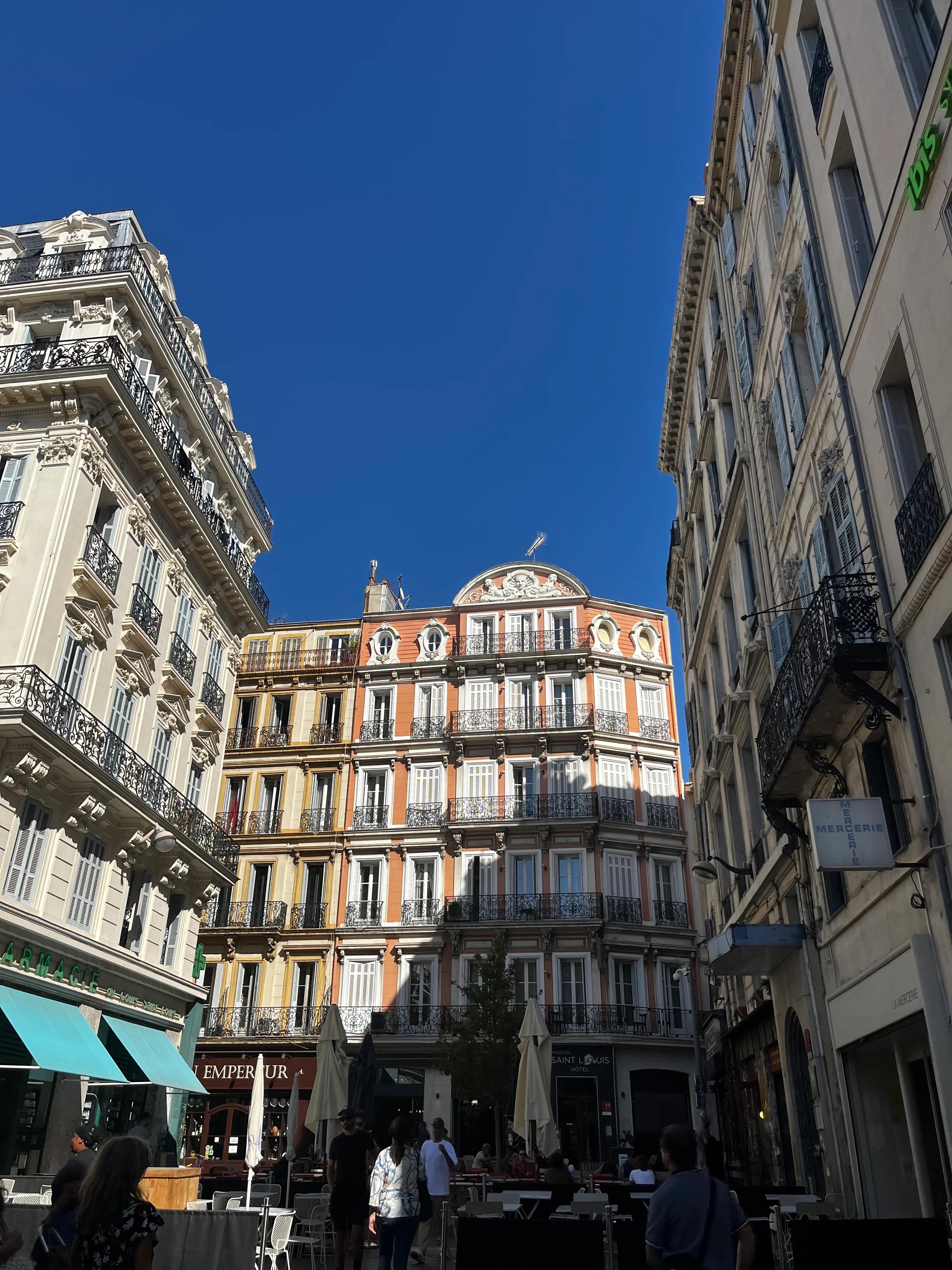 Vibrant Marseille street with ornate buildings against blue sky. People walking and seated at outdoor cafés in the sunny foreground.