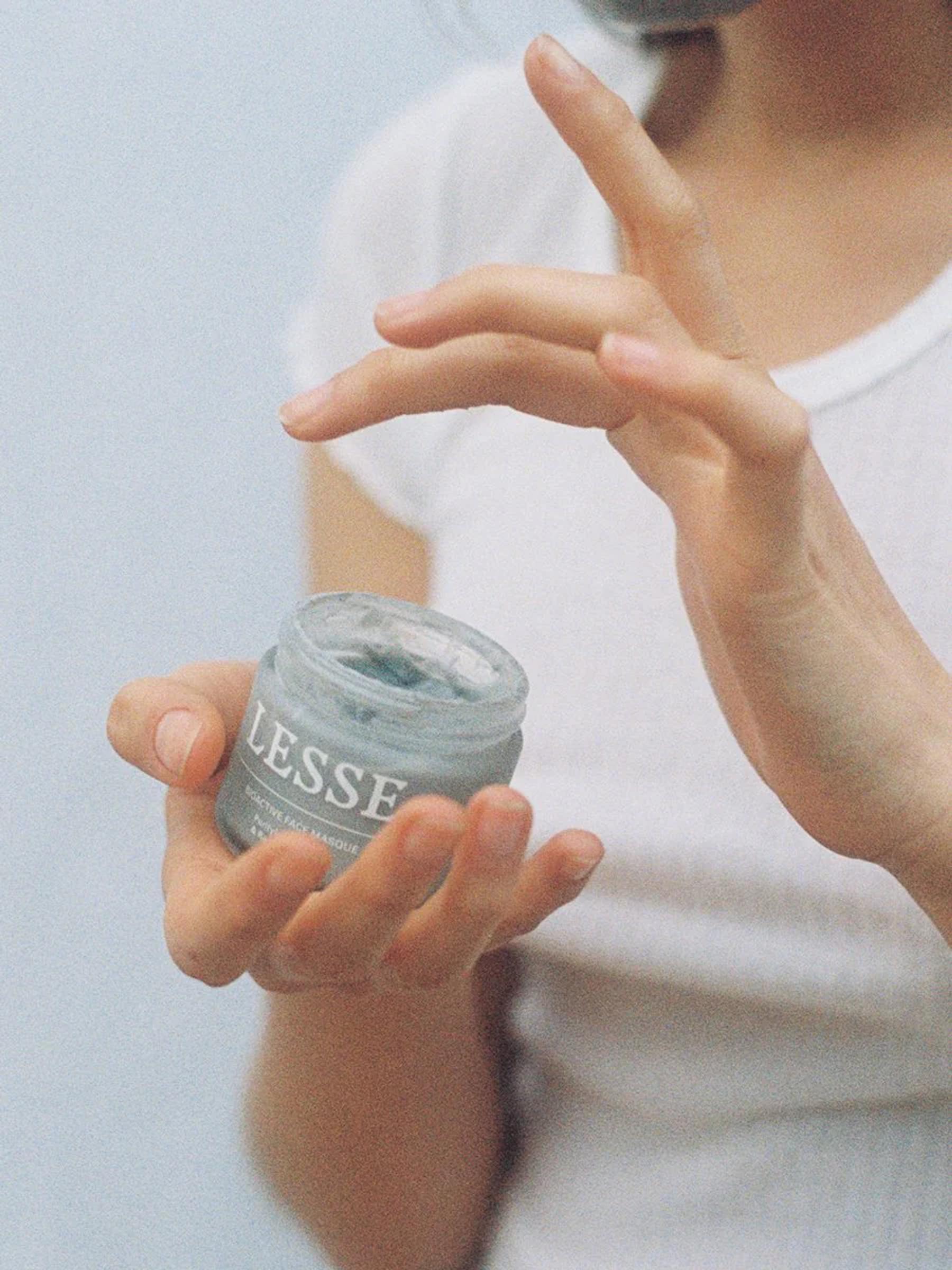 Person in white shirt dips finger into open LESSE cream jar. Soft, blurred background highlights product.