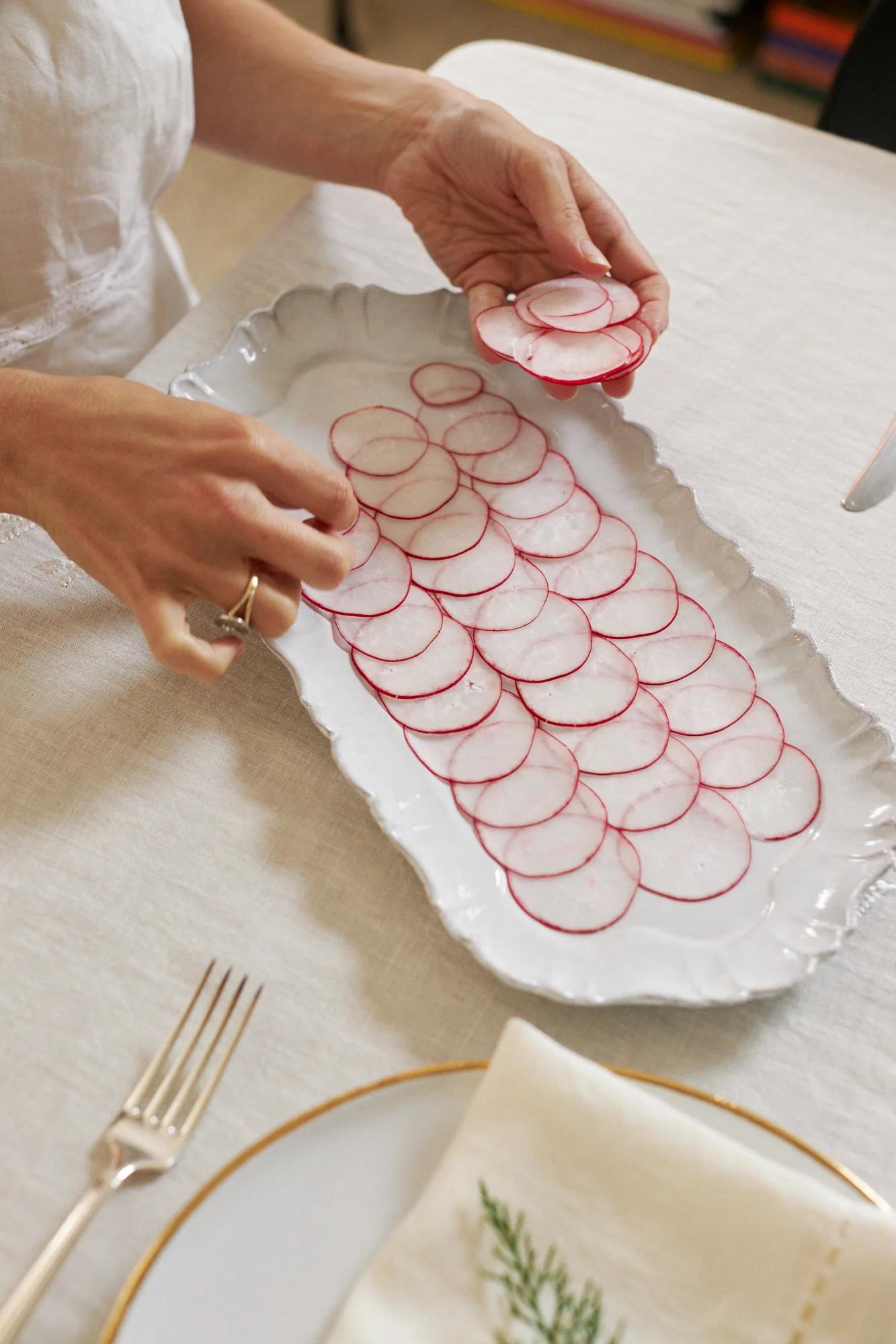 Someone arranges sliced radishes on a scalloped platter. A gold-rimmed plate, fork, and napkin with greenery are on the light tablecloth.