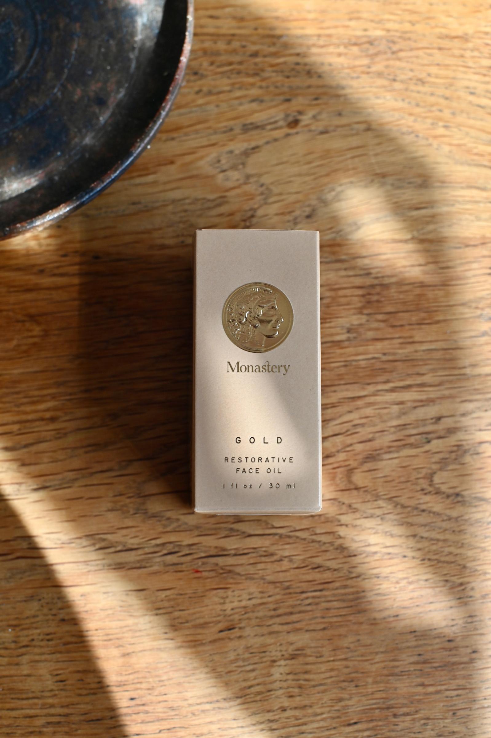 Monastery Gold botanical oil serum bottle with silver cap against neutral background. Amber oil visible, another bottle blurred in foreground.
