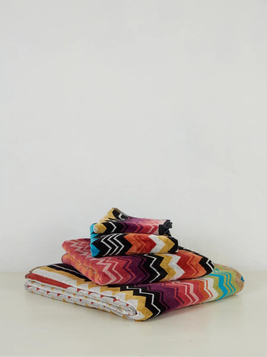 A stack of folded, colourful textiles with zigzag designs. The luxurious velour fabrics feature various hues including red, yellow, black, blue, and white.