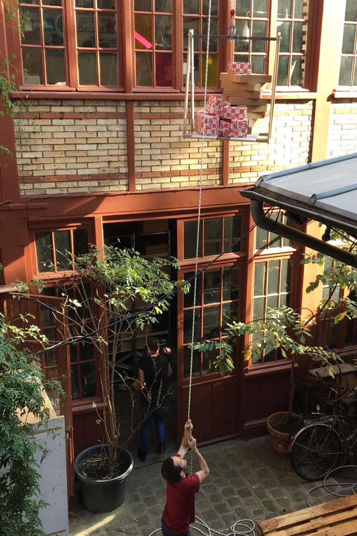 Person lifting packages on platform in courtyard. Brick walls, plants, bicycle visible. Another person seen through doorway.