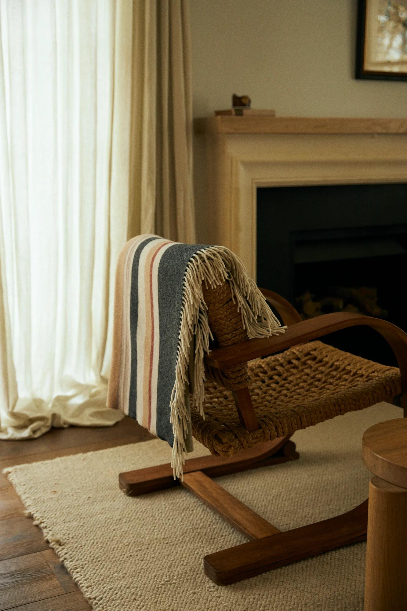 Sunlit room with woven chair, striped blanket. Black fireplace, wooden mantel. Sheer curtains on window.