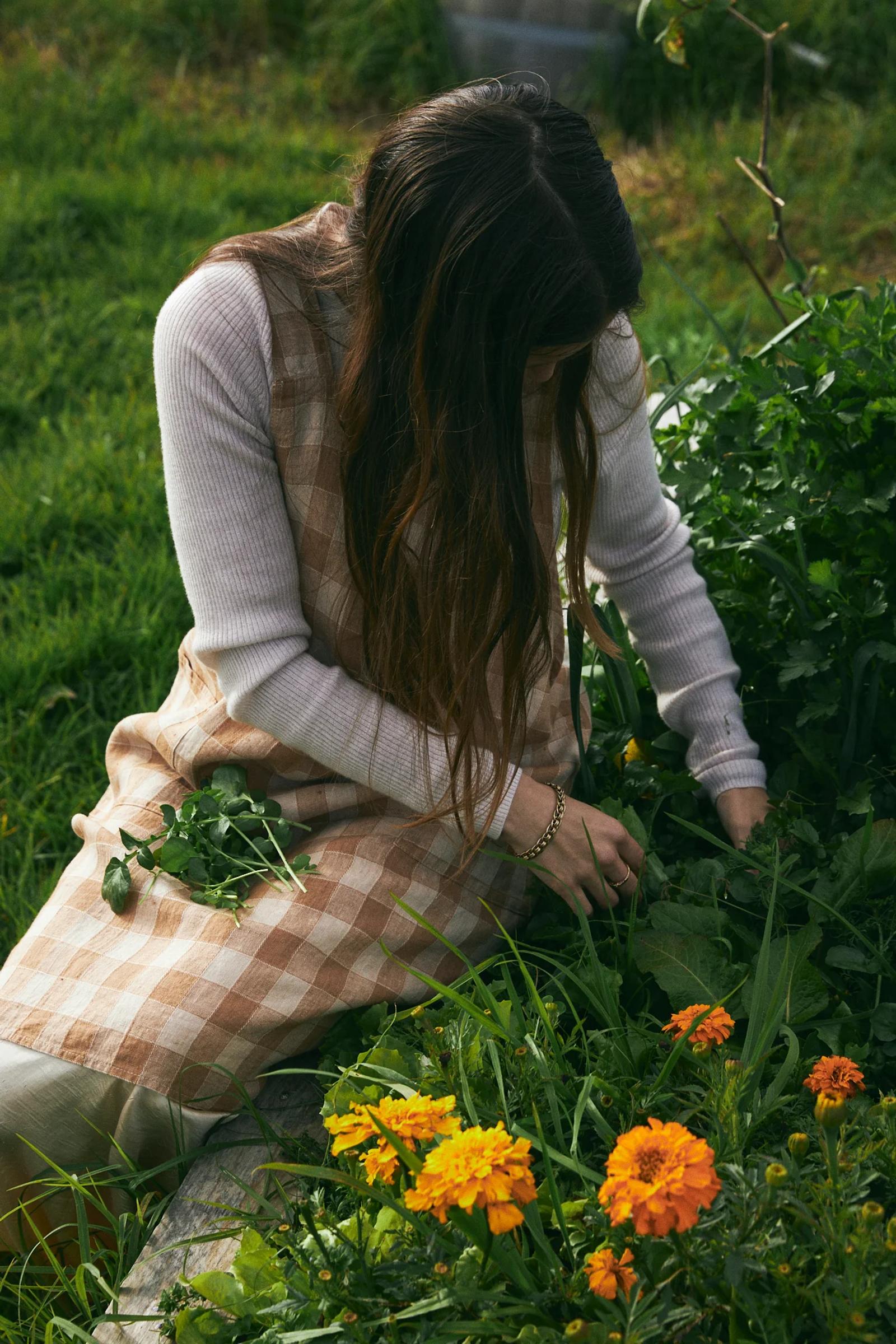 Person in checkered apron and white shirt gardening. Surrounded by greenery and yellow flowers.