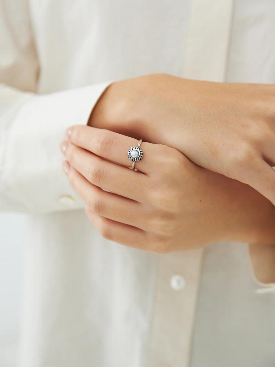 Close-up of hands showing engagement ring with round-cut gemstone. Person wears light-coloured, long-sleeved shirt.