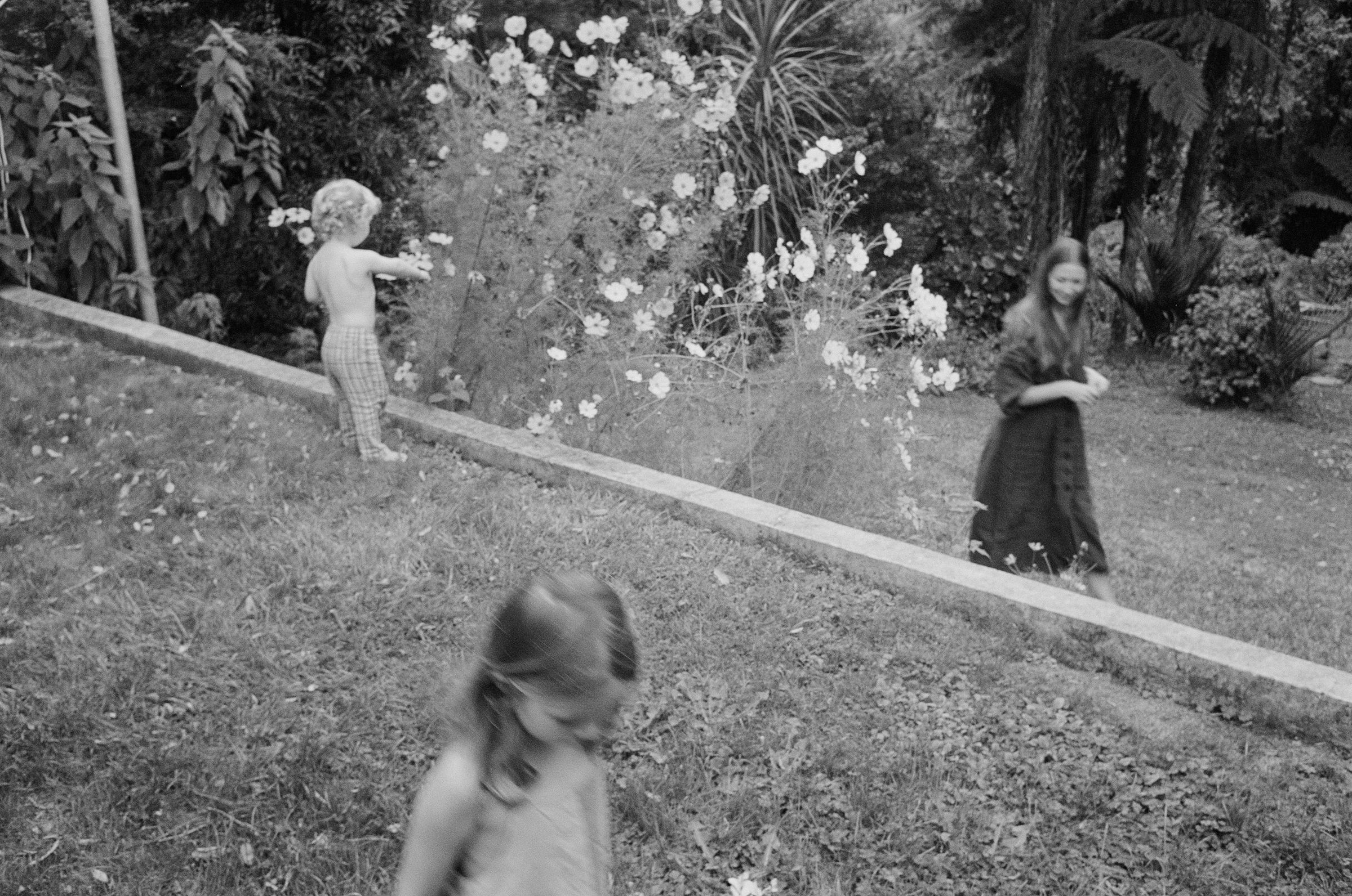 Black and white photo of three children outdoors. One shirtless near flowers, one walking, one climbing slope.