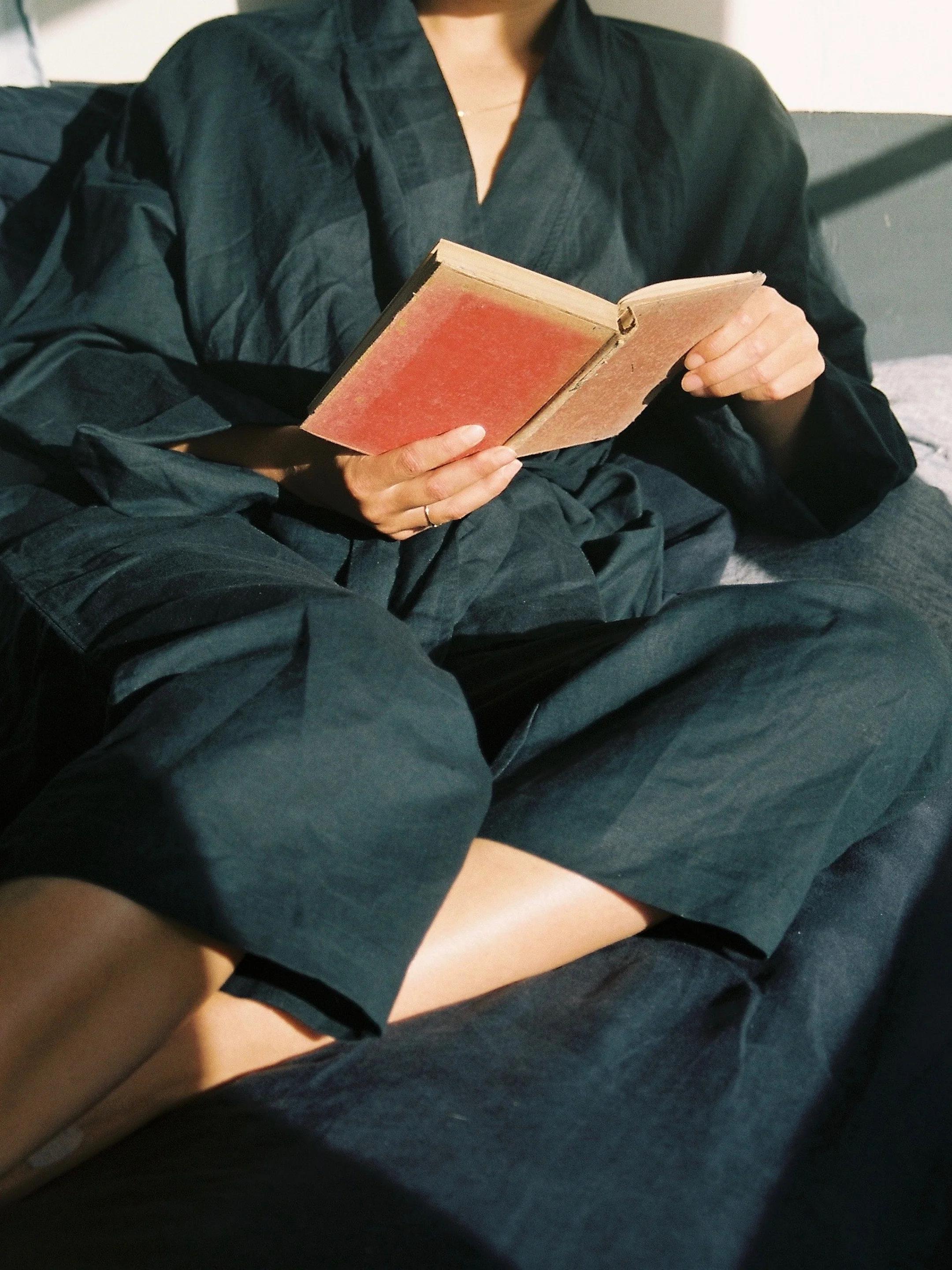 Person in dark sleepwear reads red book. Sunlight highlights outfit and book. Blurred background.