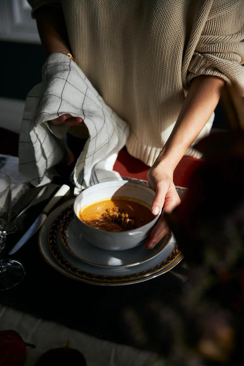 A person in a knitted sweater sets a bowl of pumpkin soup on a dark-clothed table. The cozy setting includes plates, cutlery, and a wine glass.