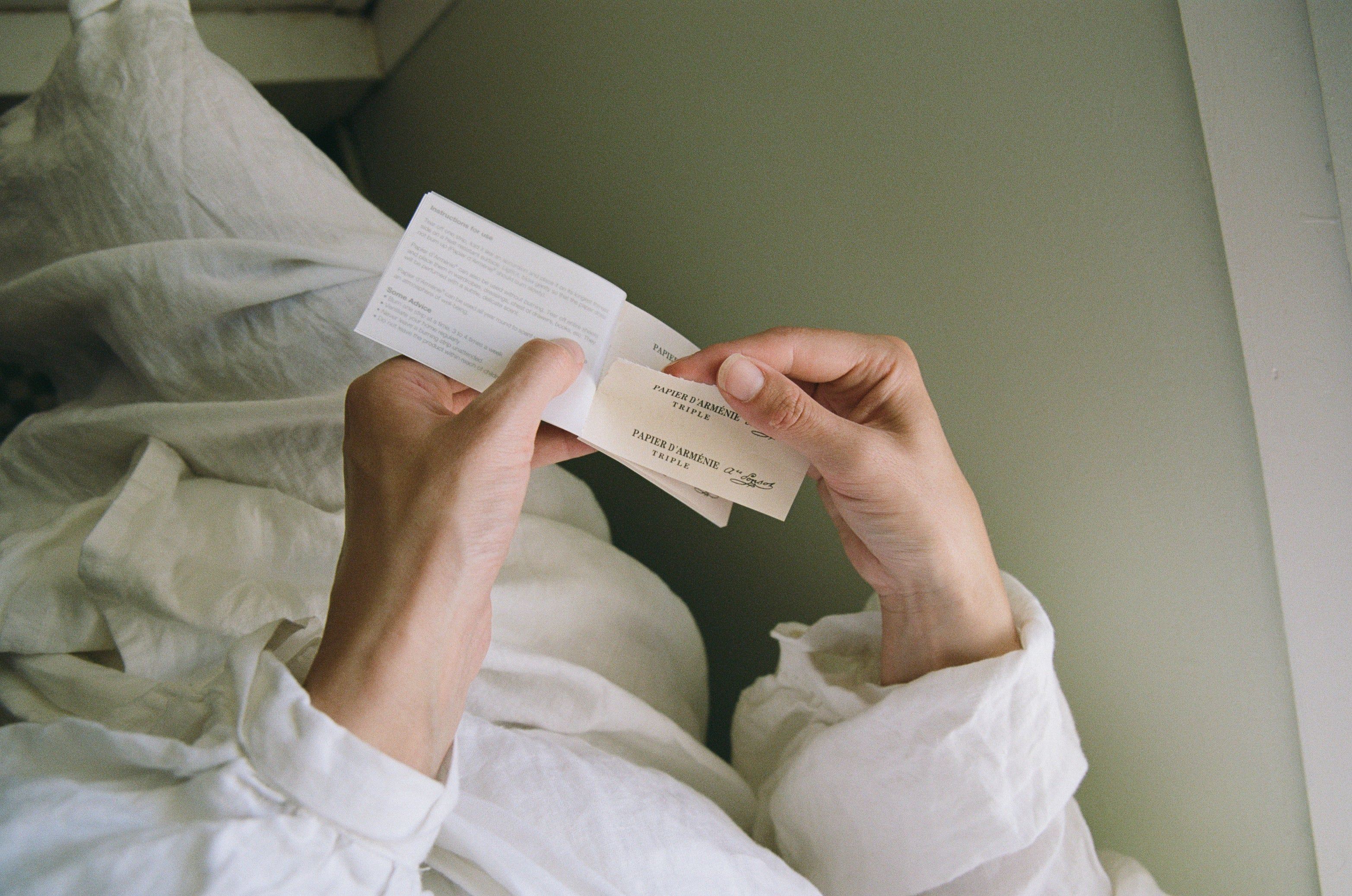 A person in white cotton reads a small white booklet, tearing out a perforated page. The light green wall background and the scene evoke an air of old-world tranquility.