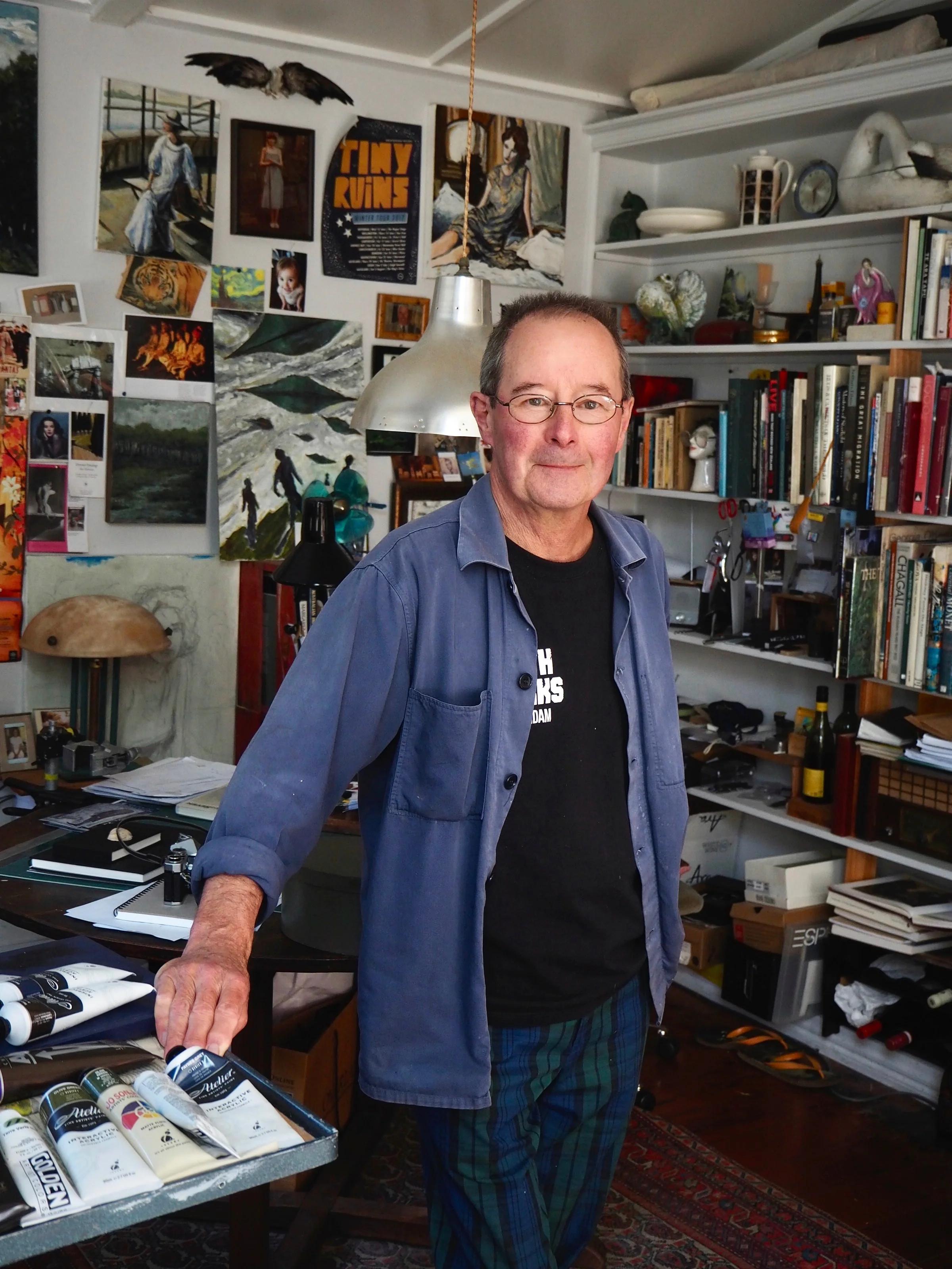 Man in blue shirt and plaid pants in art-filled studio. Max Thomson's works visible. Rests arm on paint-covered table.