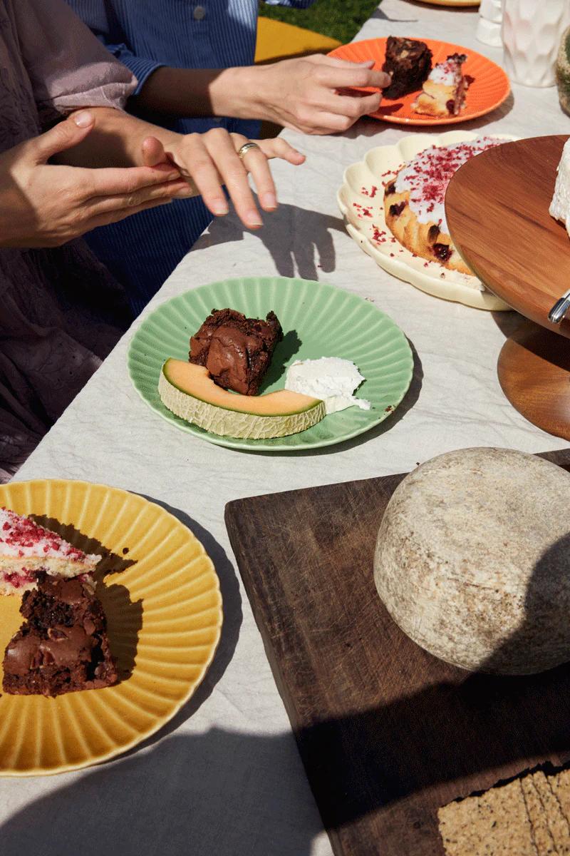 Outdoor table with colourful desserts, cakes, and brownies. Cheese wheel on wooden board. Two hands visible in conversation. Bright, sunny setting for sweet tooths.