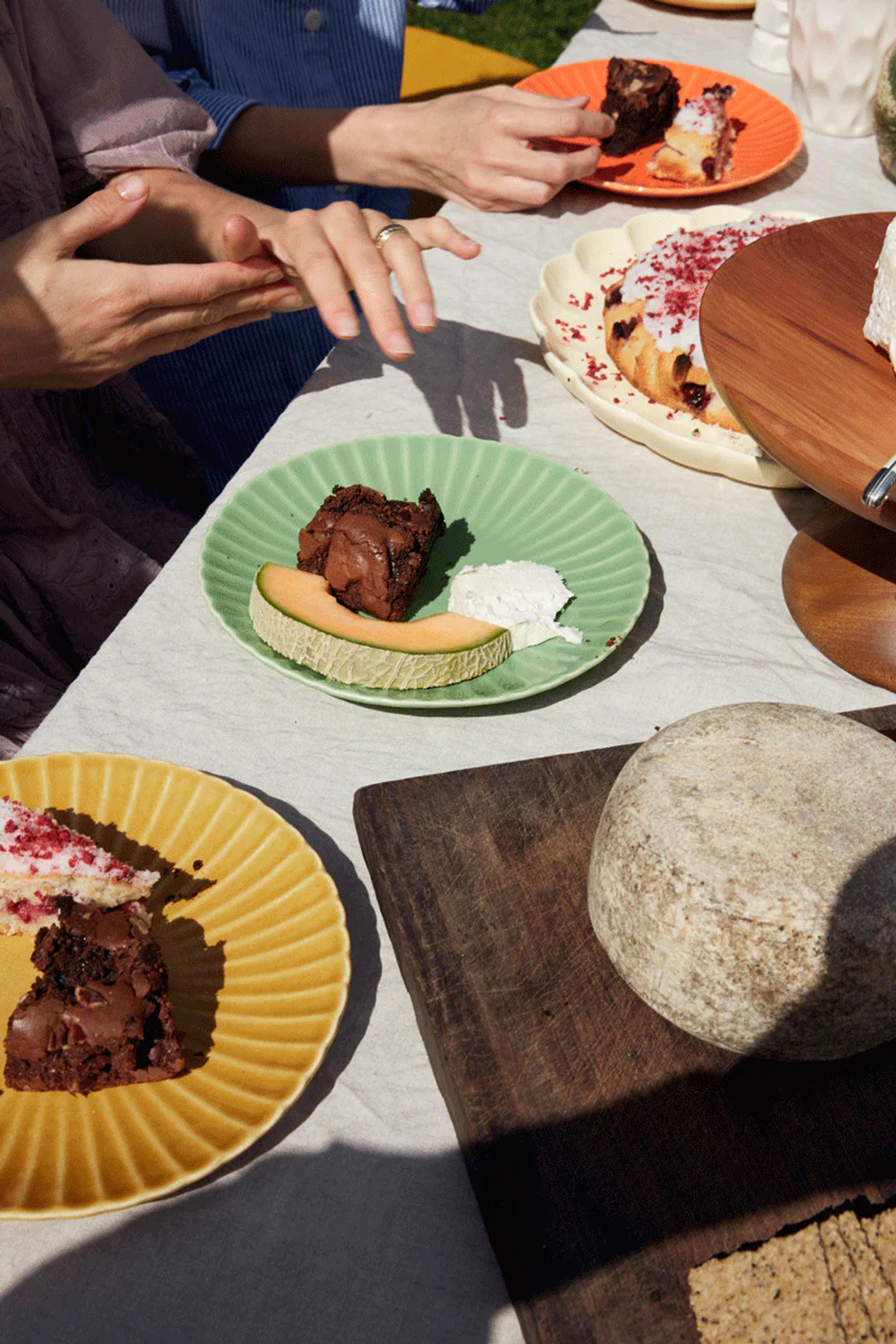 Outdoor table with colourful desserts, cakes, and brownies. Cheese wheel on wooden board. Two hands visible in conversation. Bright, sunny setting for sweet tooths.
